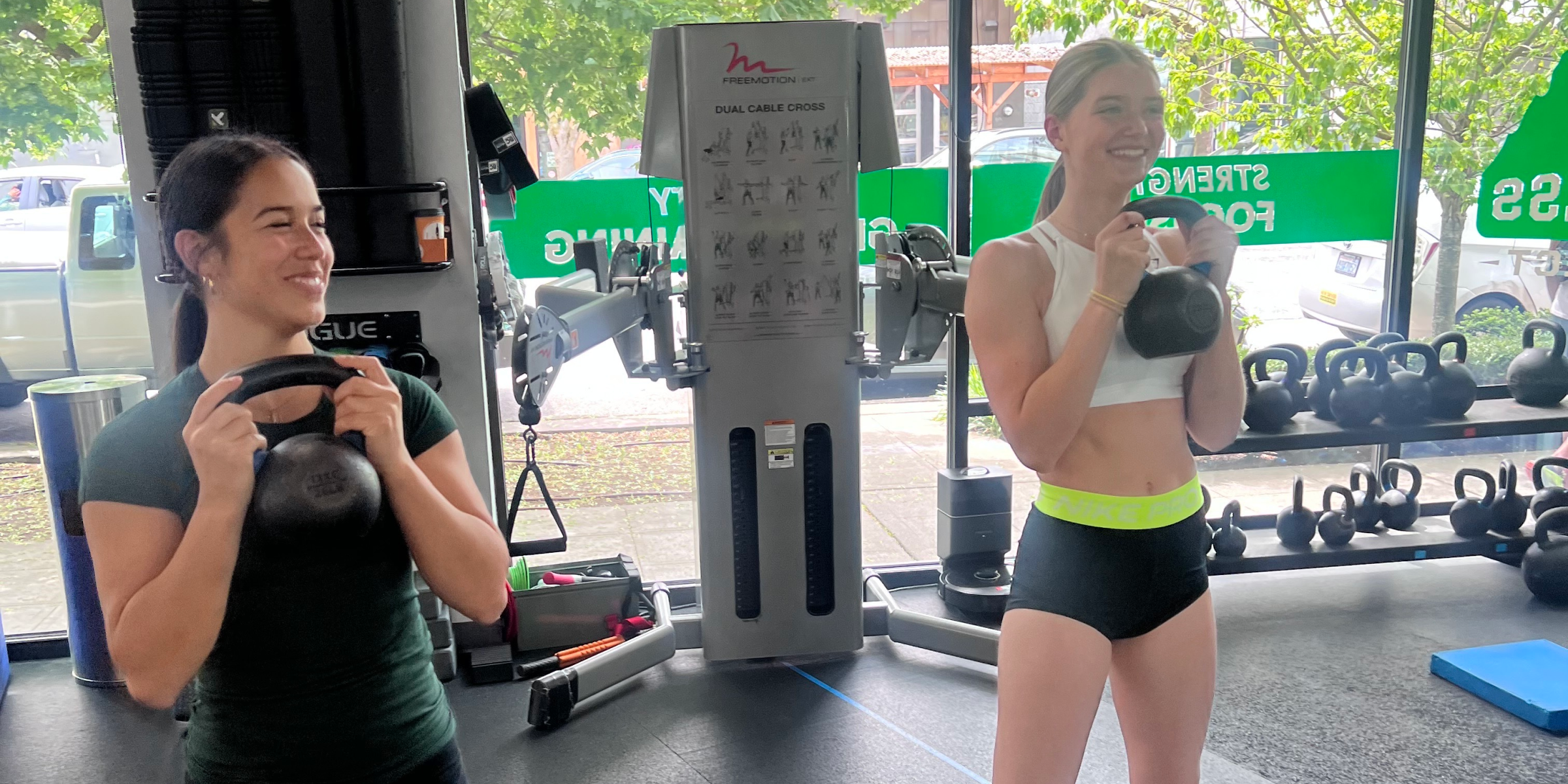 Two women getting Personal Training smiling and holding kettlebells inside a Seattle Fitness studio, with workout equipment and large windows showing parked cars and trees outside.