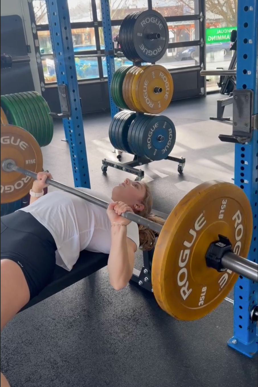 A Personal Training client lying on a bench, lifting a barbell with weight plates, and do a bench press in a Personal training studio with weight racks and colorful plates in the background.
