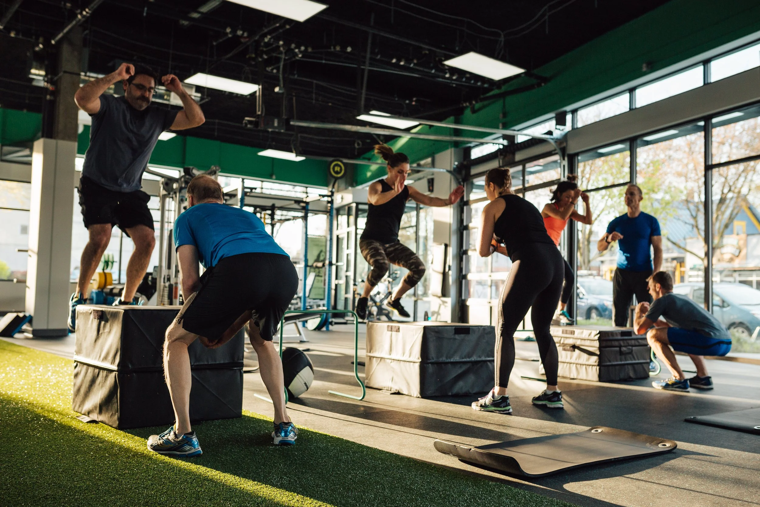 People working out with a trainer in a gym with large windows, sunlight coming in, and various exercise equipment.