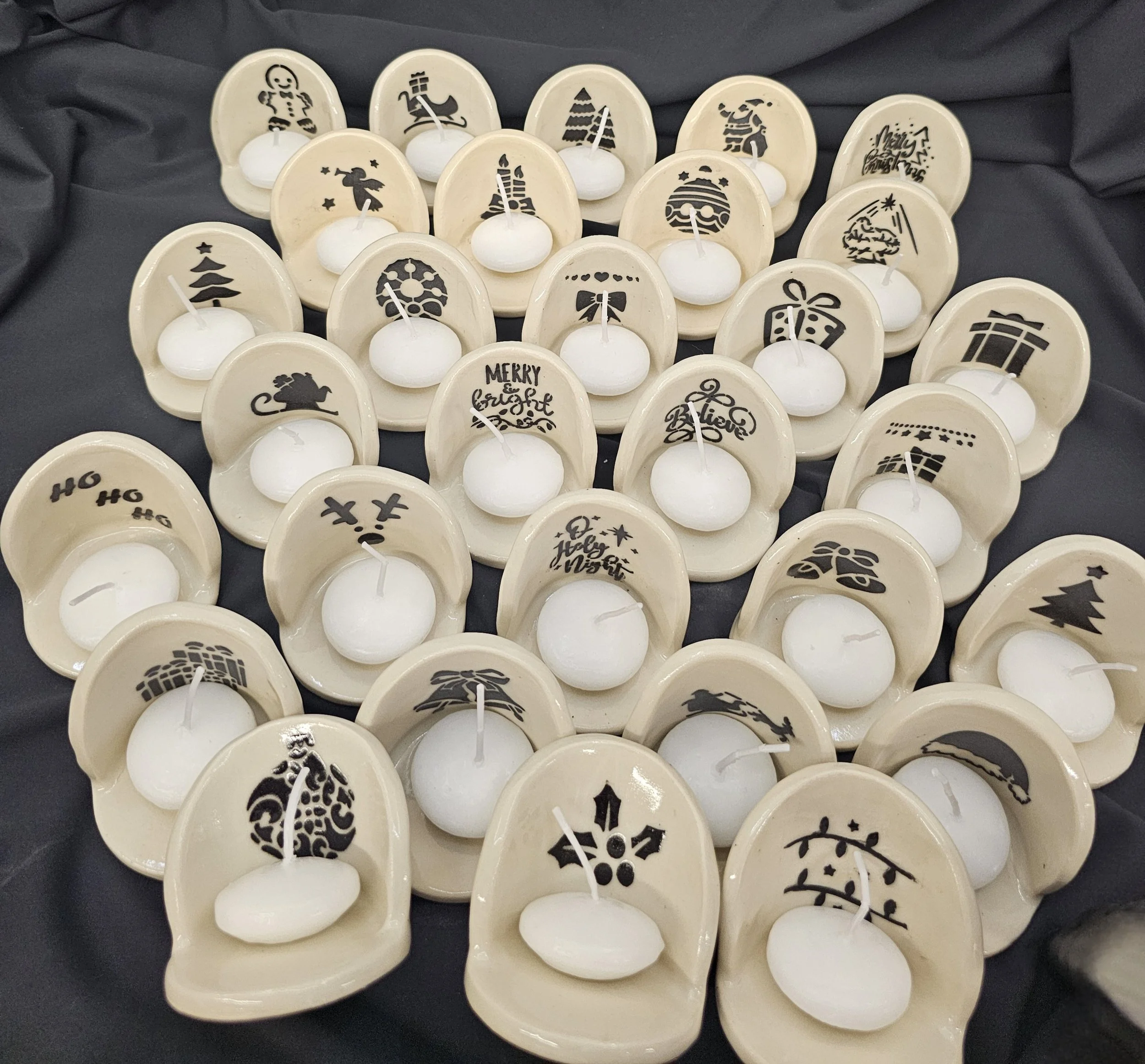 Multiple white candles placed in ceramic holders with black holiday-themed designs on a dark cloth background.