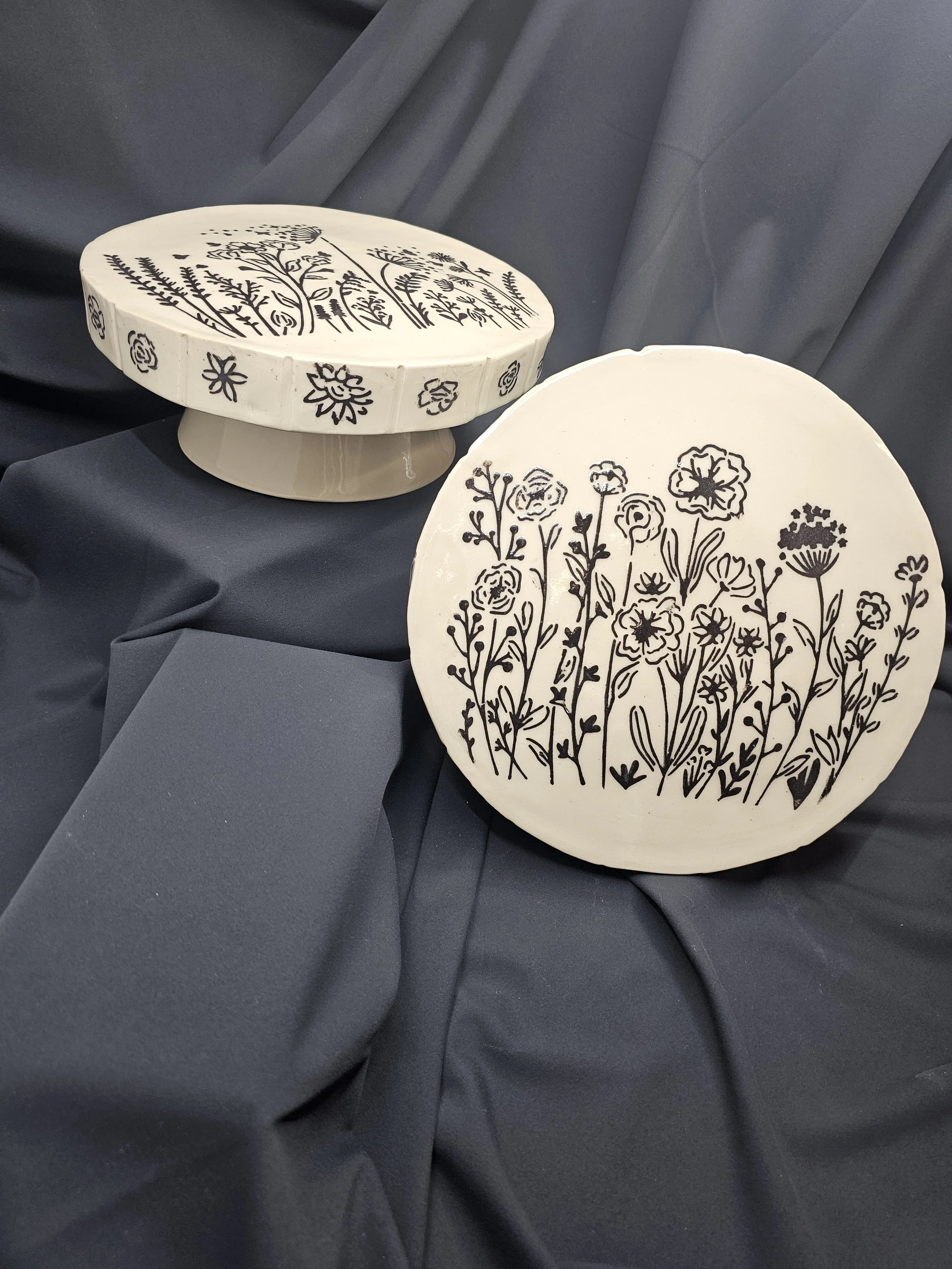 Round ceramic plates with black floral and plant patterns, placed on dark fabric. One plate is upright, the other tilted.
