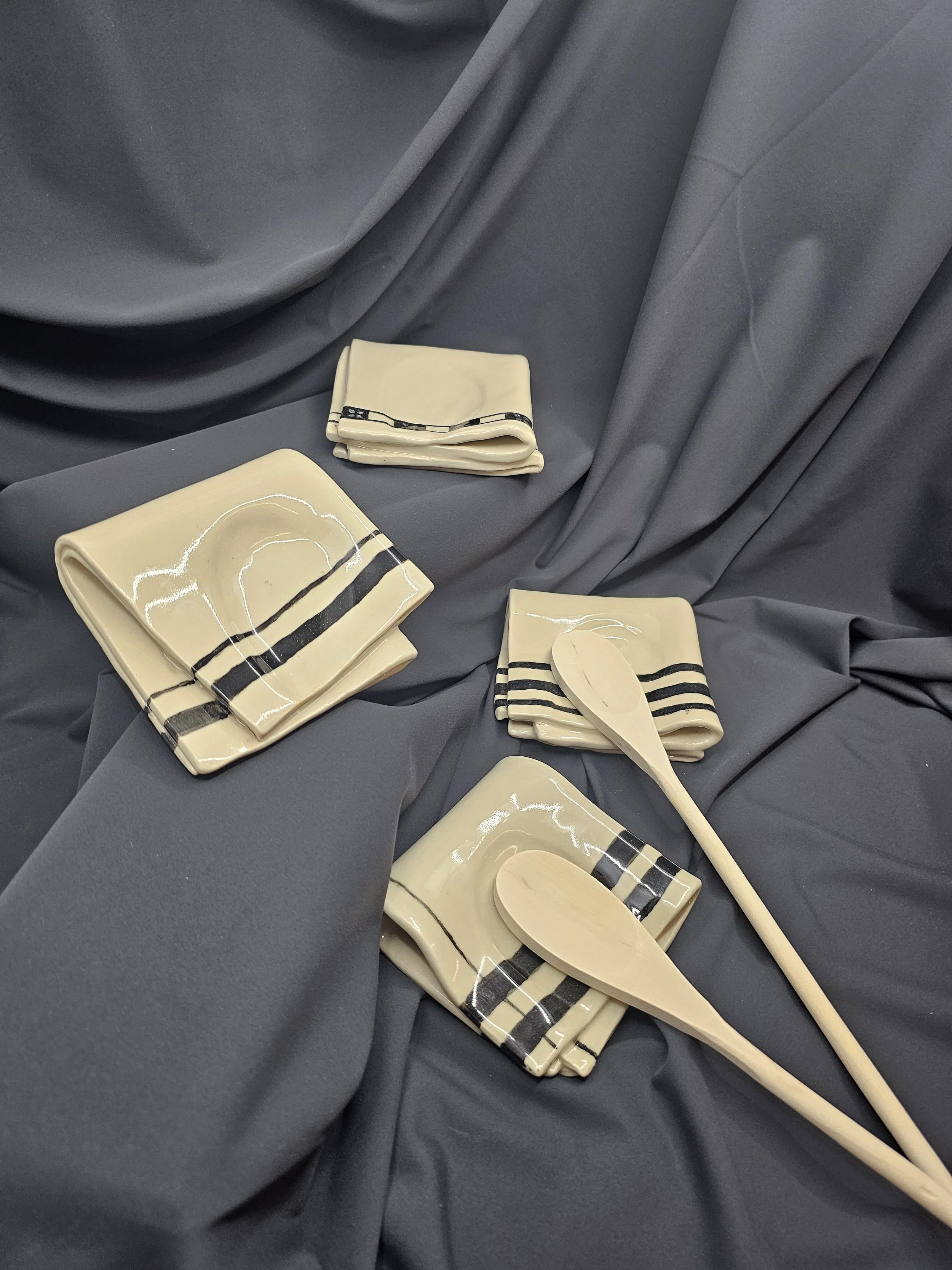 Ceramic dishware set including square plates and spoons with black stripe designs, arranged on gray fabric background.