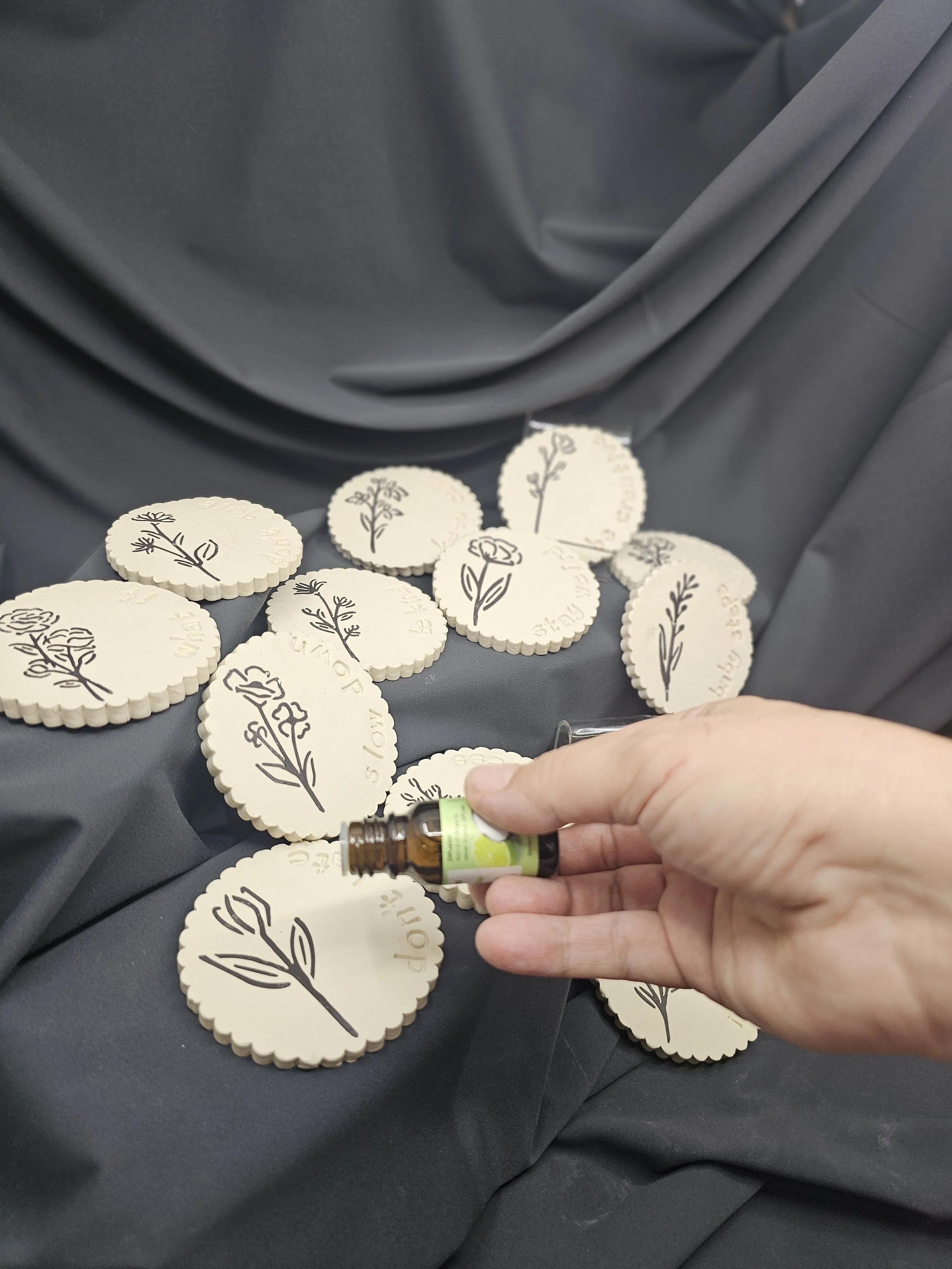 A hand holding a small bottle of essential oil over round cream-colored tokens with black floral designs and text, placed on a dark fabric background.