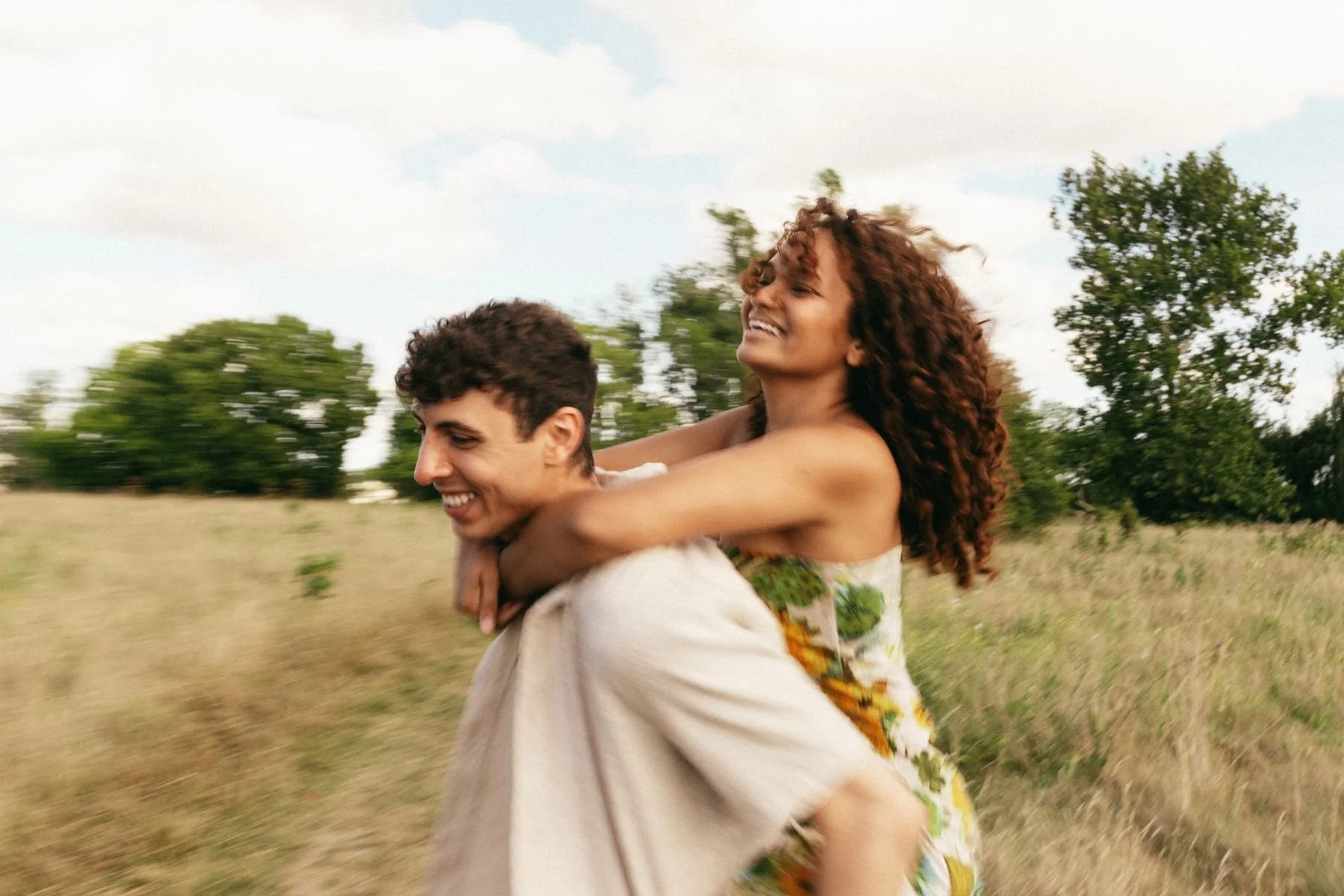 A joyful young woman with curly hair being carried on a man's back through a grassy field with trees and a partly cloudy sky in the background.