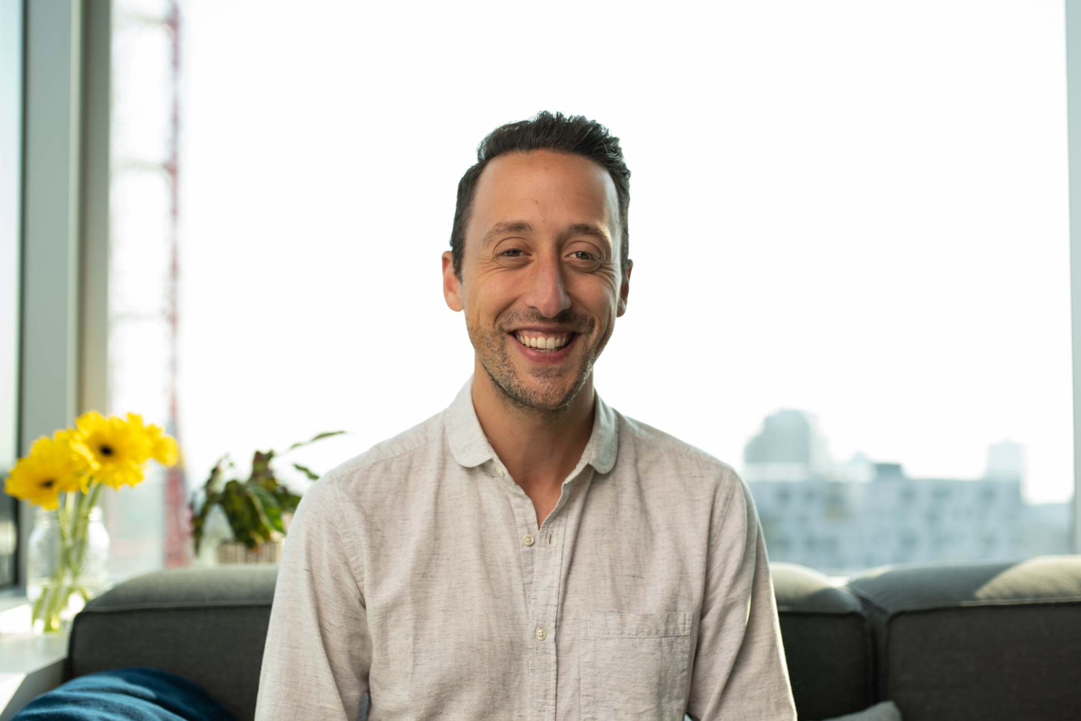 Smiling man with dark hair and light-colored shirt sitting on a couch in a bright room with large windows.
