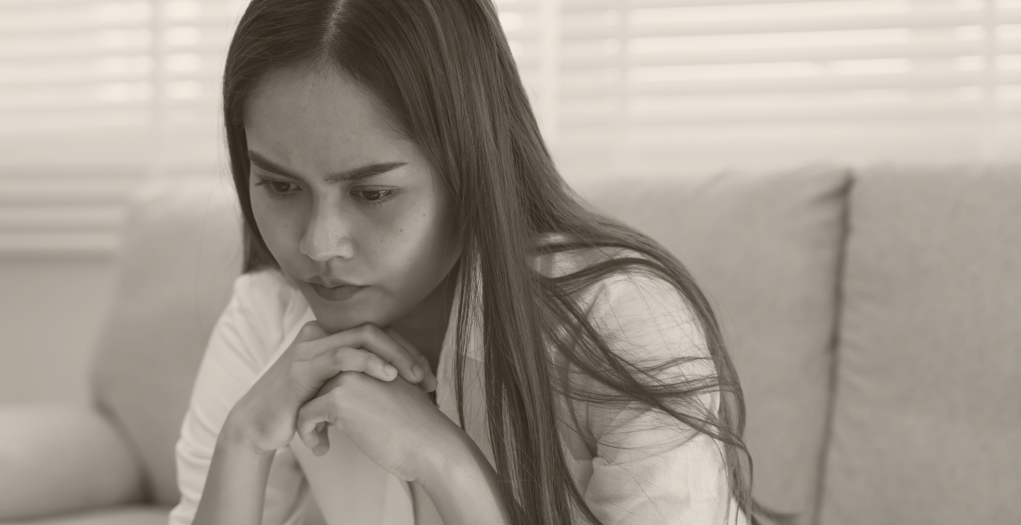 A young woman with long hair looking down thoughtfully with her hands clasped under her chin, sitting on a couch with blinds in the background.