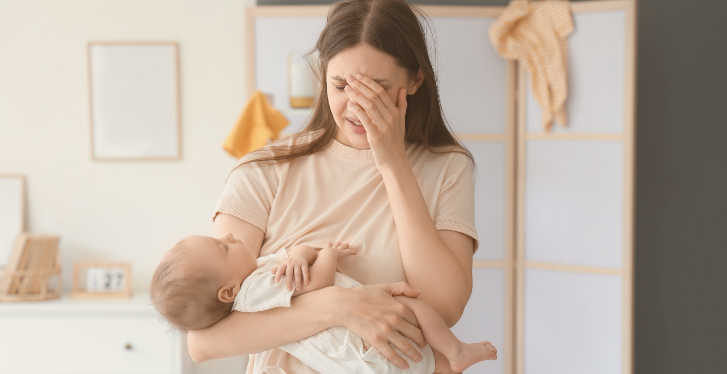 A woman holding a baby and looking distressed, with her hand covering her face.