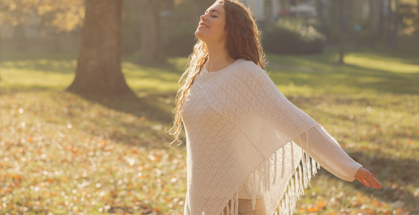 Woman with long hair enjoying the sunshine with arms outstretched in a park during fall.