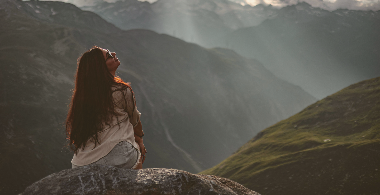 A woman sitting on a rock overlooking green mountains with mist in the background.