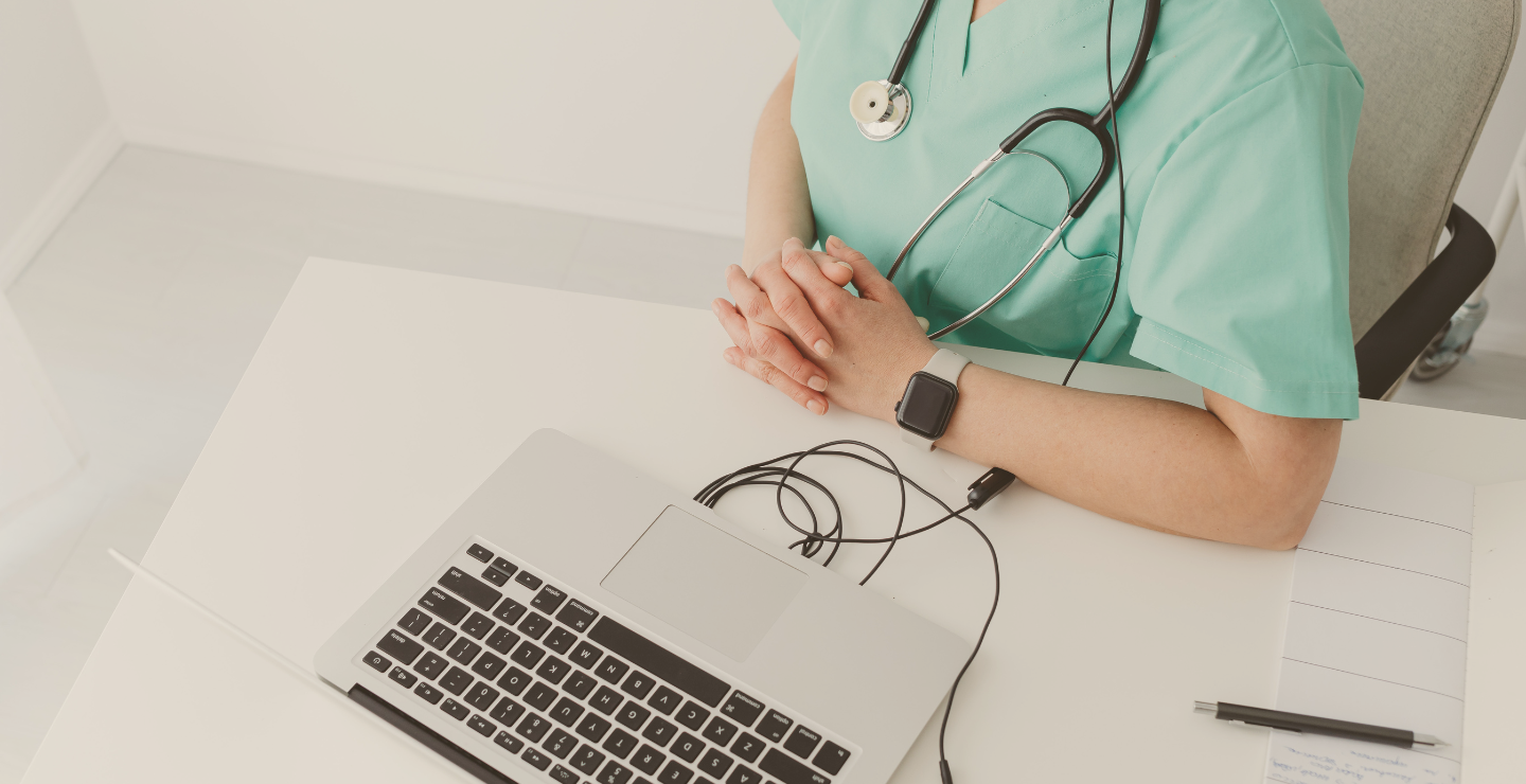 A healthcare worker in scrubs sitting at a white desk with hands clasped. A laptop, pen, notepad, and a smartwatch are on the desk. The healthcare worker wears a stethoscope around their neck and an Apple Watch on their wrist.