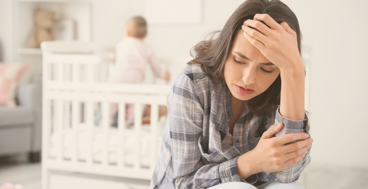 Woman with a headache, sitting on the floor near a crib, with a baby in the background.
