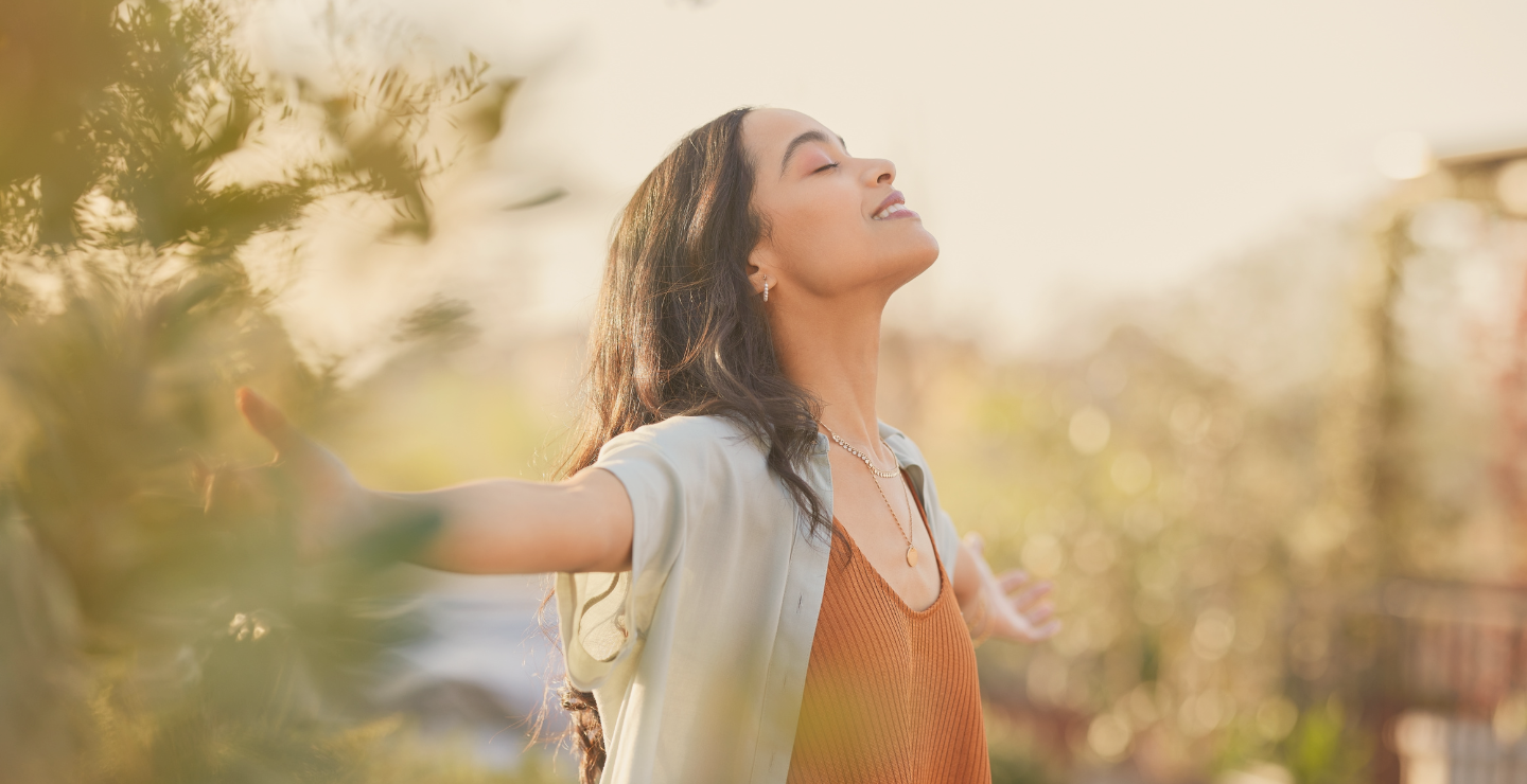 A woman with long dark hair standing outdoors with her arms outstretched and eyes closed, smiling, basking in sunlight during daytime.
