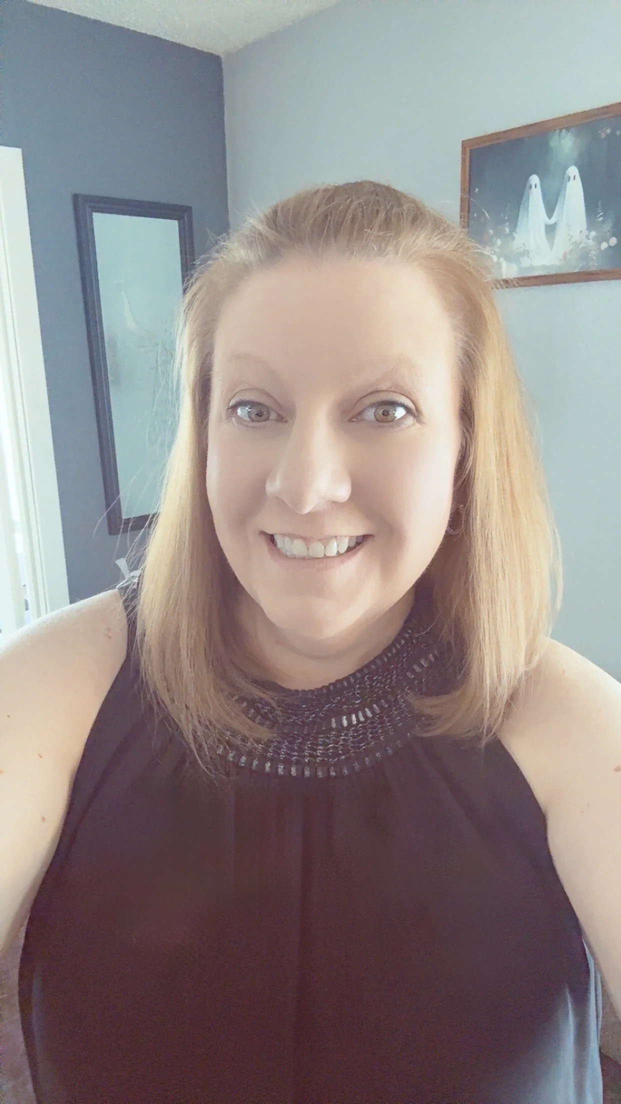 Close-up selfie of a smiling woman with shoulder-length light brown hair, wearing a black sleeveless top with a decorative collar, in a room with blue walls and framed artwork in the background.