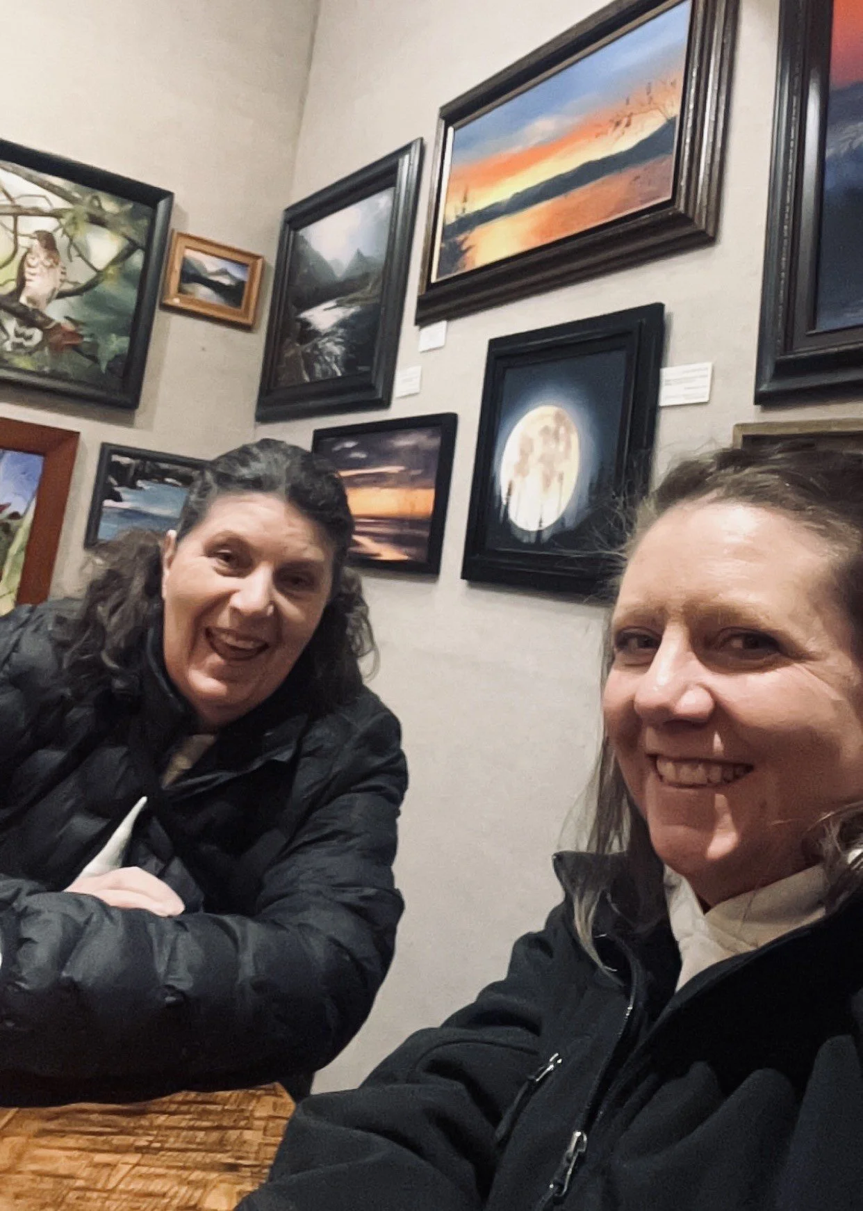 Two women smiling and sitting at a table in an art gallery, with framed landscape paintings on the wall behind them.