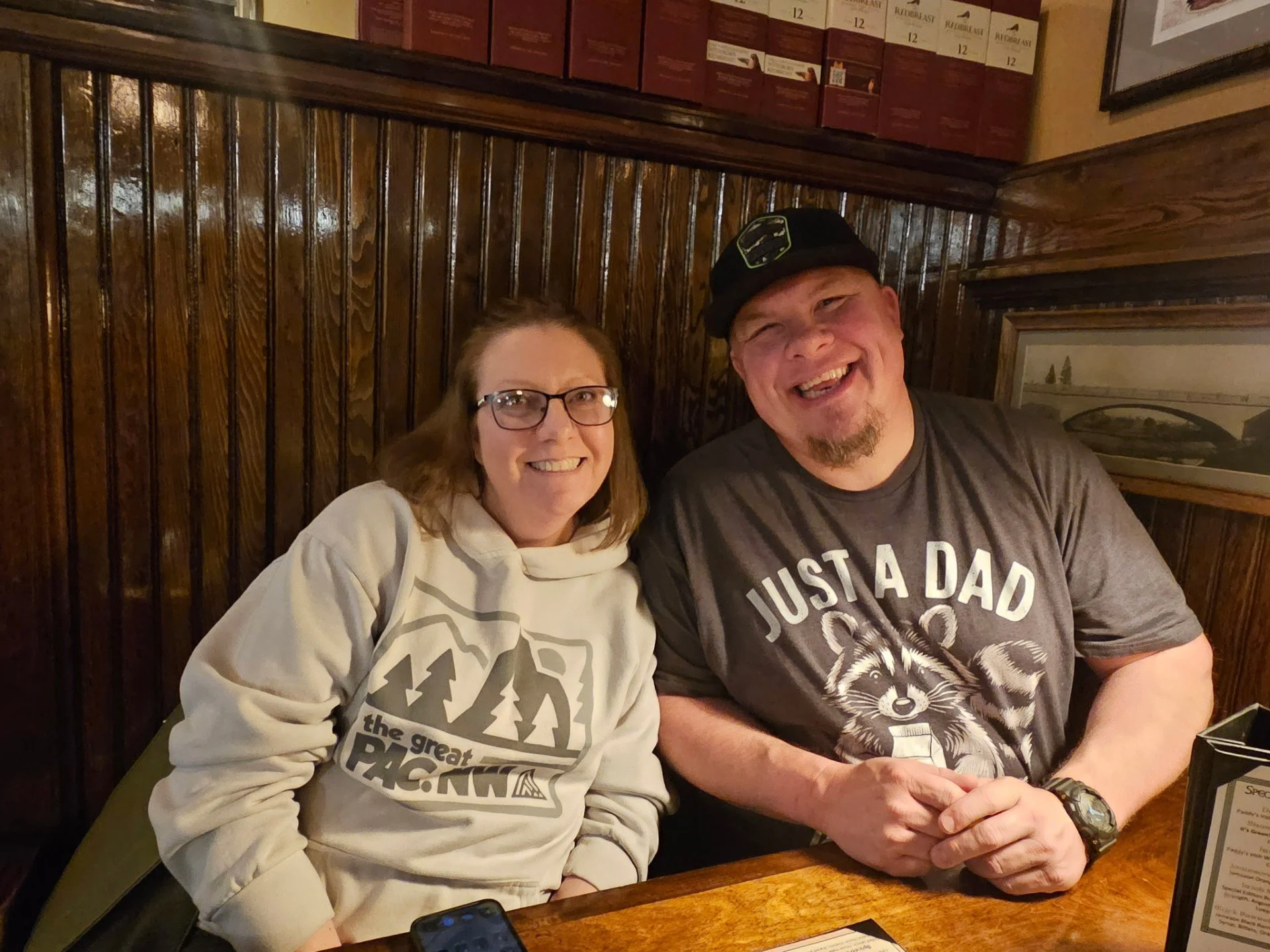 A woman and a man sitting together at a table in a restaurant, smiling for the camera. The woman has shoulder-length hair, glasses, and is wearing a light-colored hoodie. The man has a beard, is wearing a black cap and a gray T-shirt with a raccoon and the words "Just a Dad." They are seated against a dark wood-paneled wall, with framed pictures above them and a menu stand on the table.