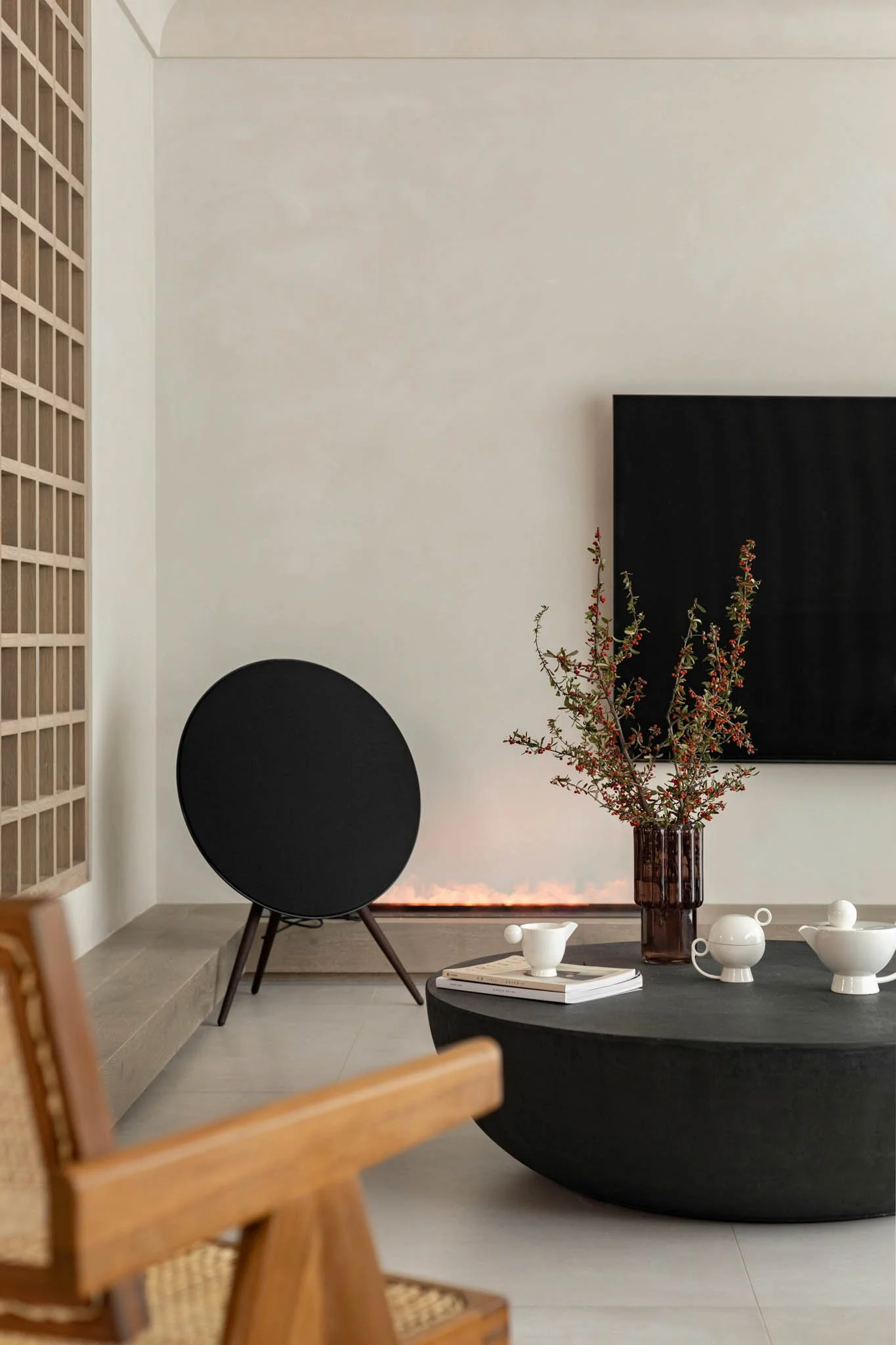 Modern living room with a black speaker, a large black framed artwork, a tall brown vase with red and green foliage, white decorative items on a black coffee table, and a wooden chair in the foreground.