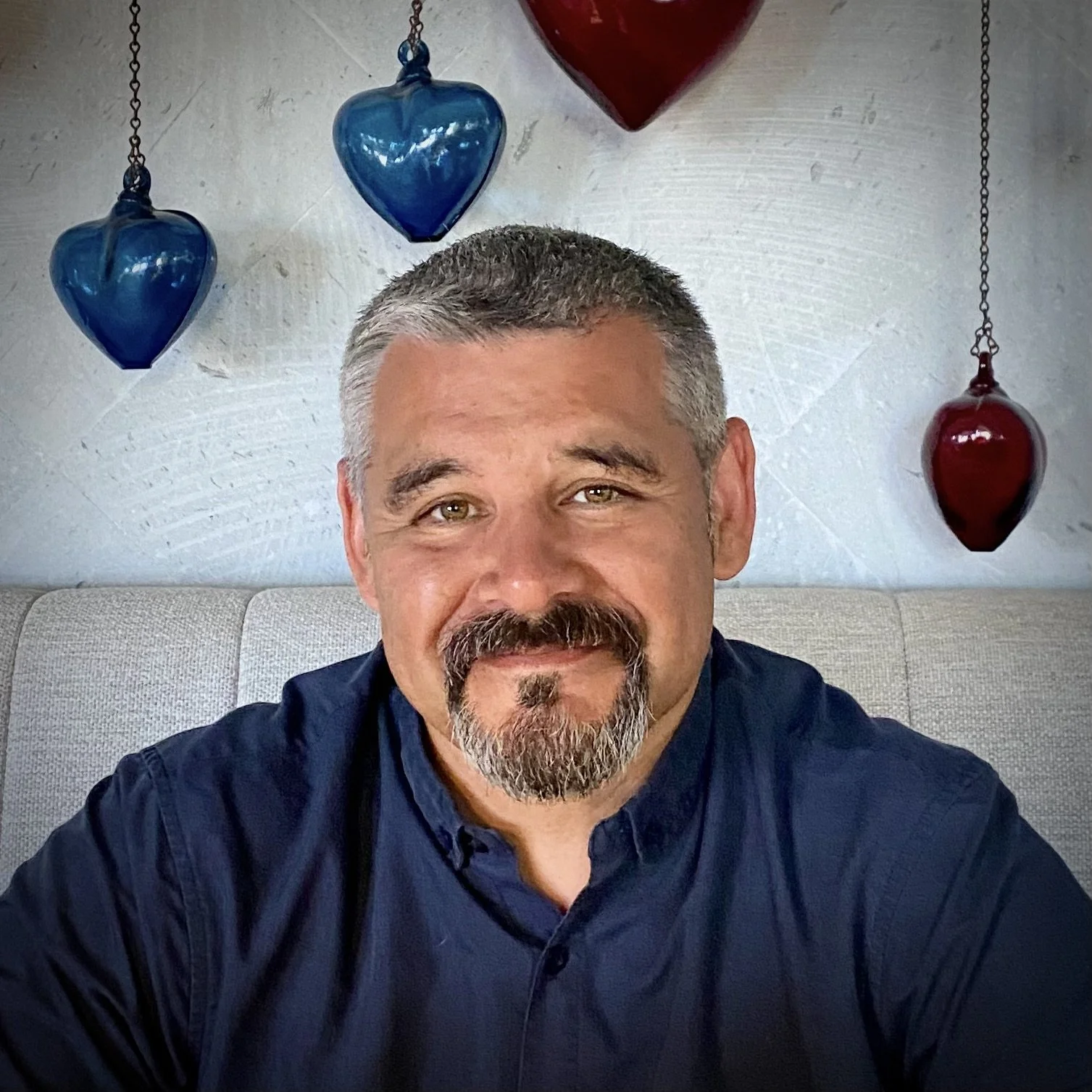 A middle-aged man with short gray hair, a mustache, and beard, smiling while sitting on a light-colored sofa. Hanging on the wall behind him are three blue and two red glass heart-shaped decorations.