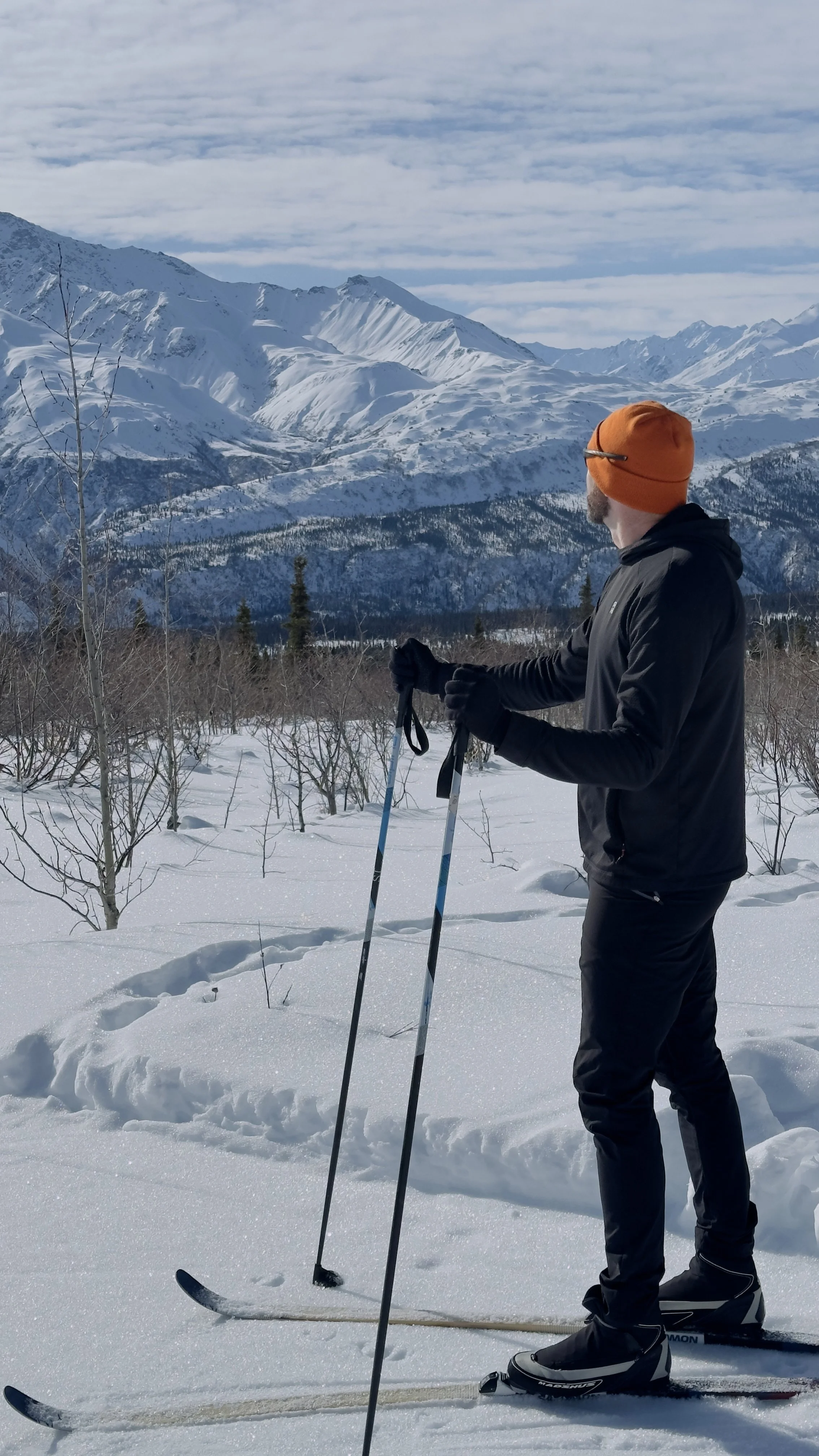 A man in black outdoor clothing and an orange beanie stands on skis in snowy terrain, holding ski poles and looking towards snow-capped mountains in the distance under a partly cloudy sky.