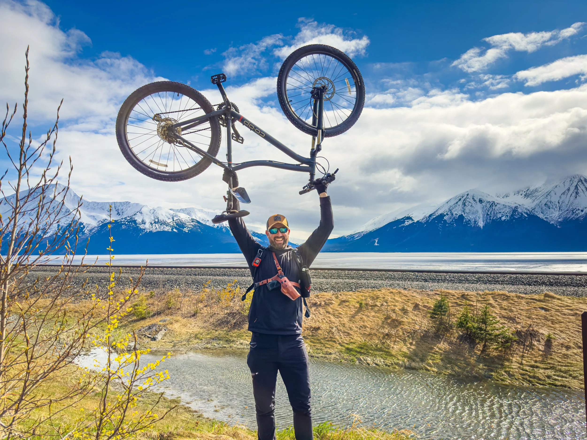 A man stands outdoors holding a mountain bike above his head with both hands. He is wearing sunglasses, a cap, and outdoor gear. The background features snow-capped mountains, a lake, and a cloudy sky.