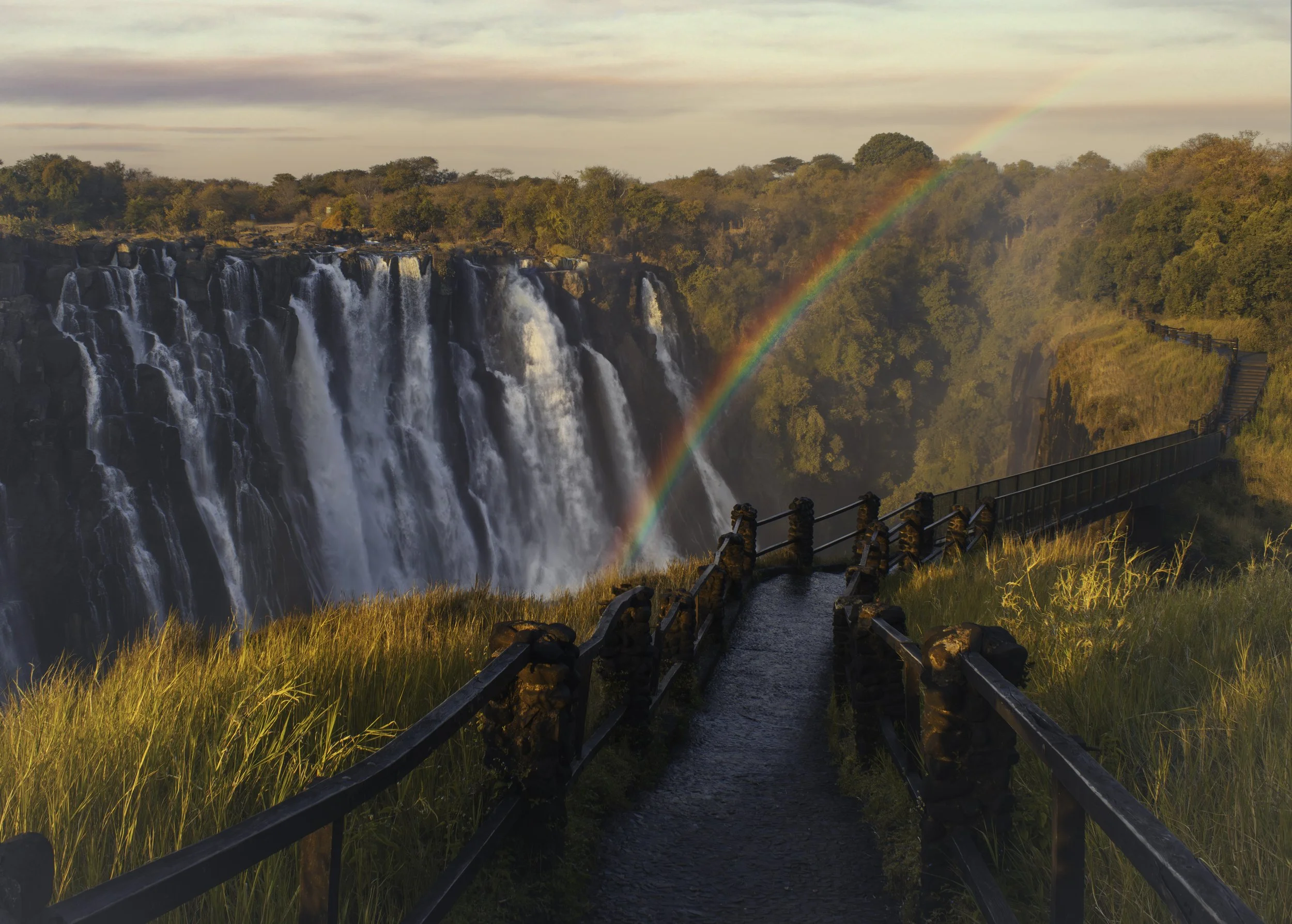 A scenic view of a waterfall with a rainbow arching above, a walkway along the edge, and lush trees in the background during sunset.