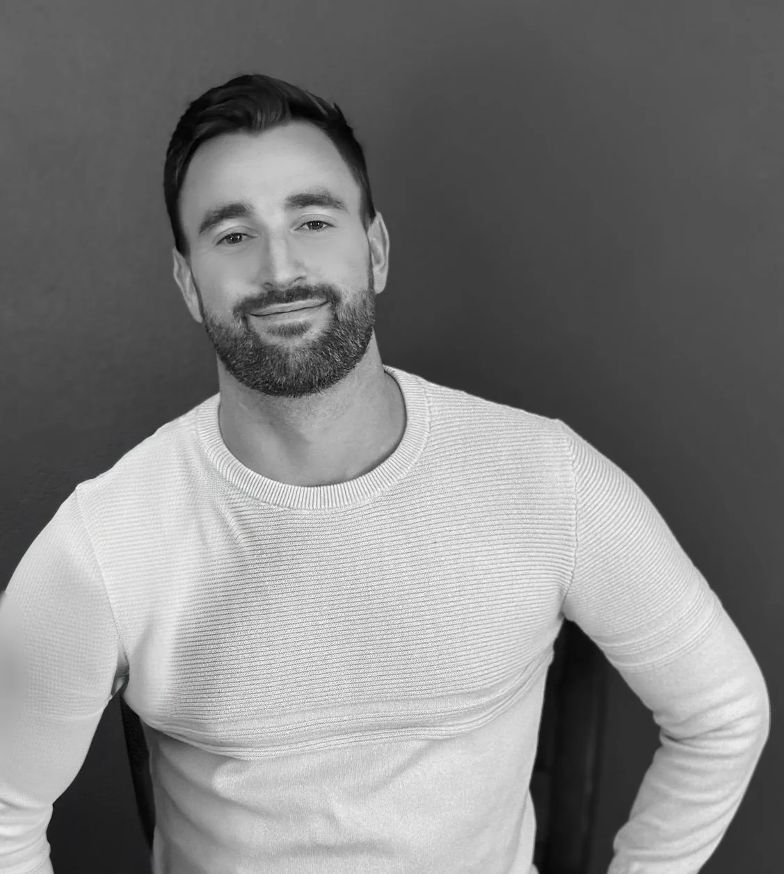 Black and white portrait of a smiling man with a beard, wearing a light-colored, ribbed sweater, standing against a plain dark background.