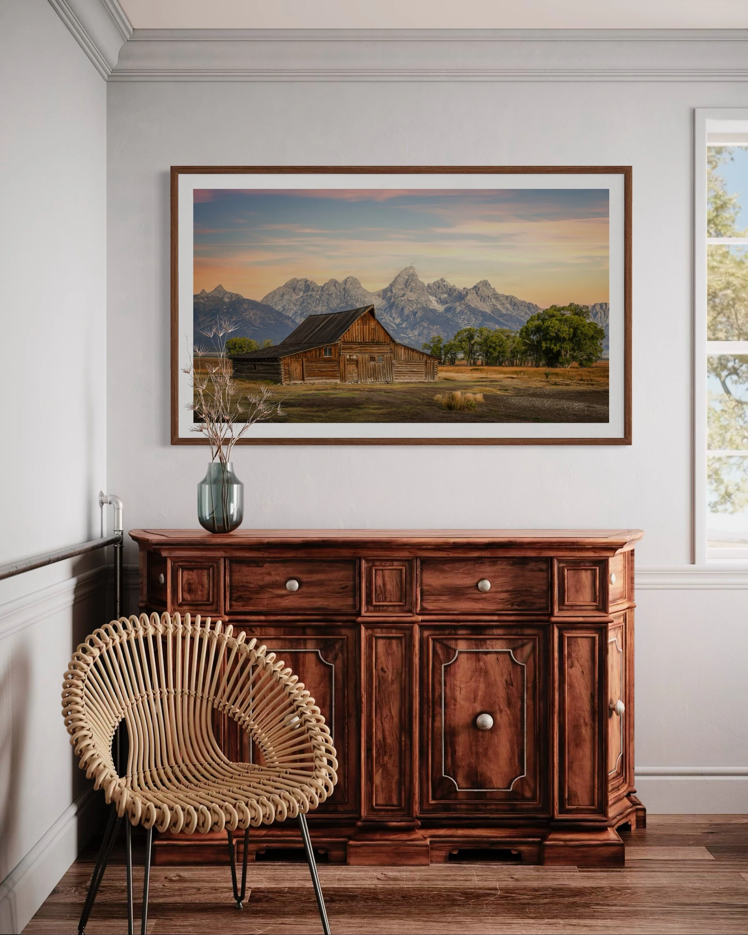 A white-walled room with a framed landscape photograph of mountains, trees, and a barn hanging above a wooden sideboard. On the sideboard is a gray vase with dried branches. A rattan chair with black metal legs is in front of the sideboard, and a window with white framing is on the right side, allowing natural light to illuminate the room.