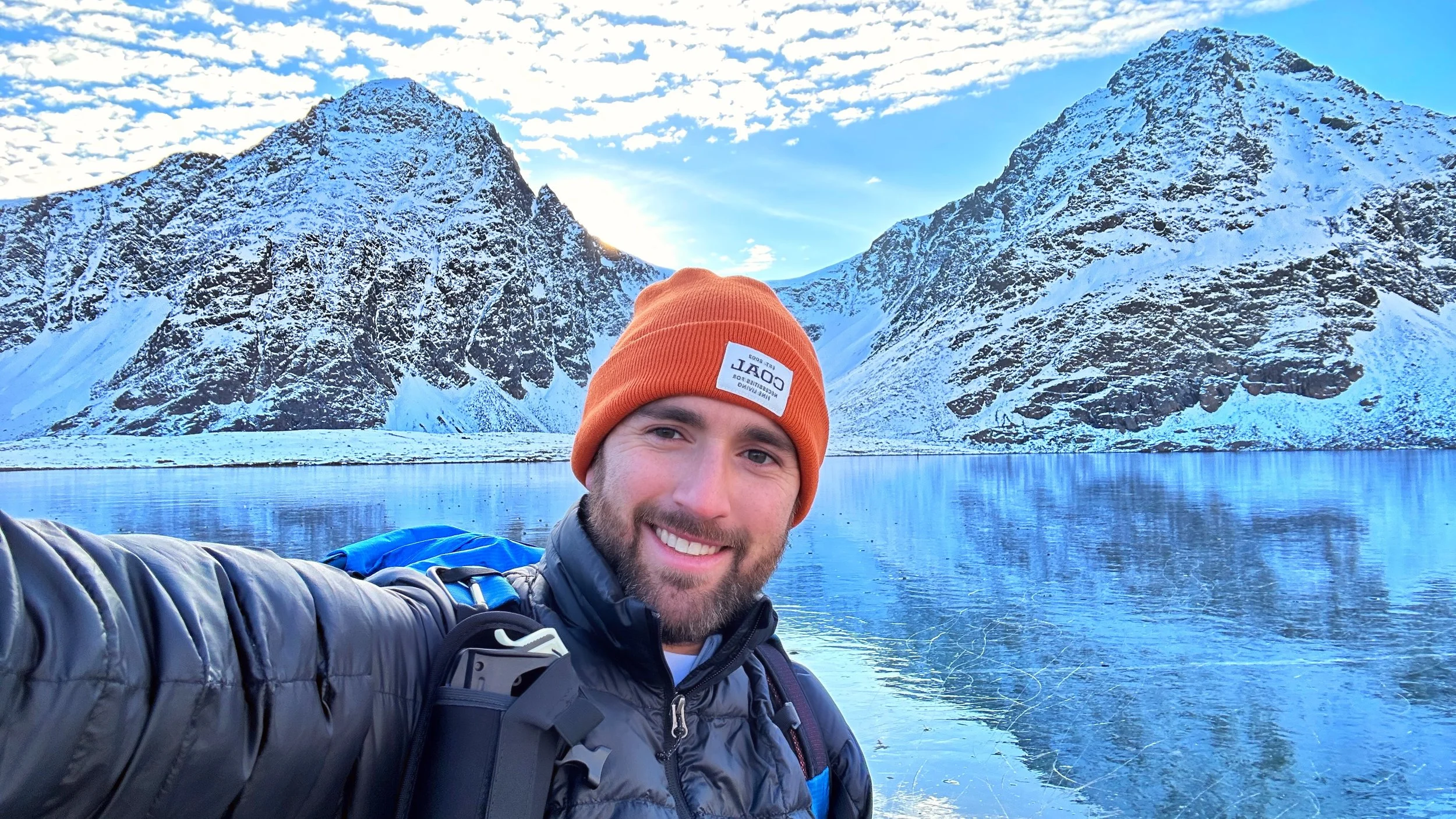 A man wearing an orange beanie and gray jacket taking a selfie near a lake with snow-covered mountains in the background.