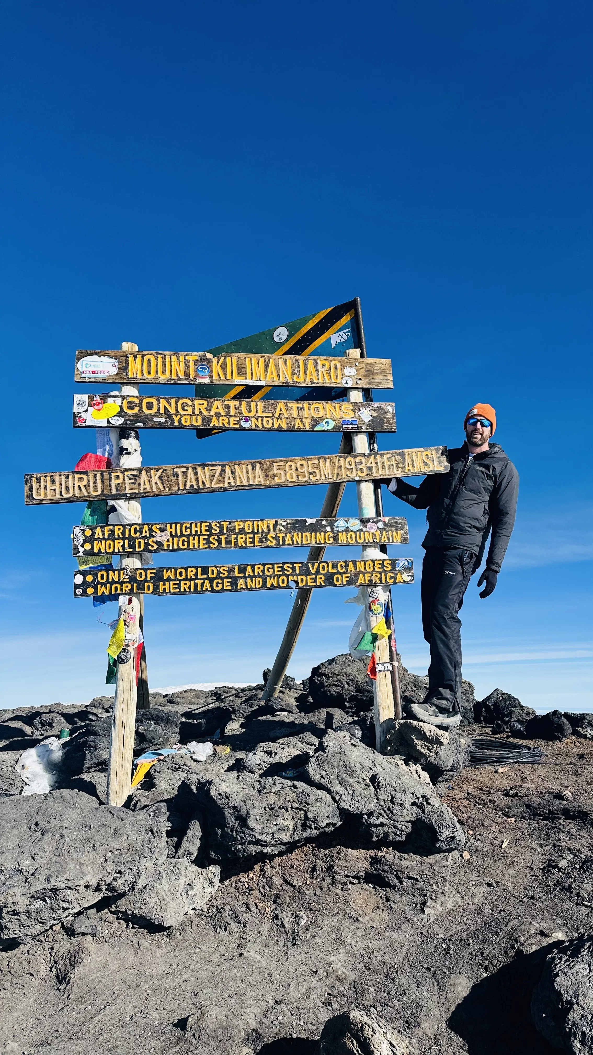 Person in black jacket, orange hat, and sunglasses standing next to a mountain summit sign at Mount Kilimanjaro, Tanzania, pointing toward the summit, with clear blue sky and rocky terrain.