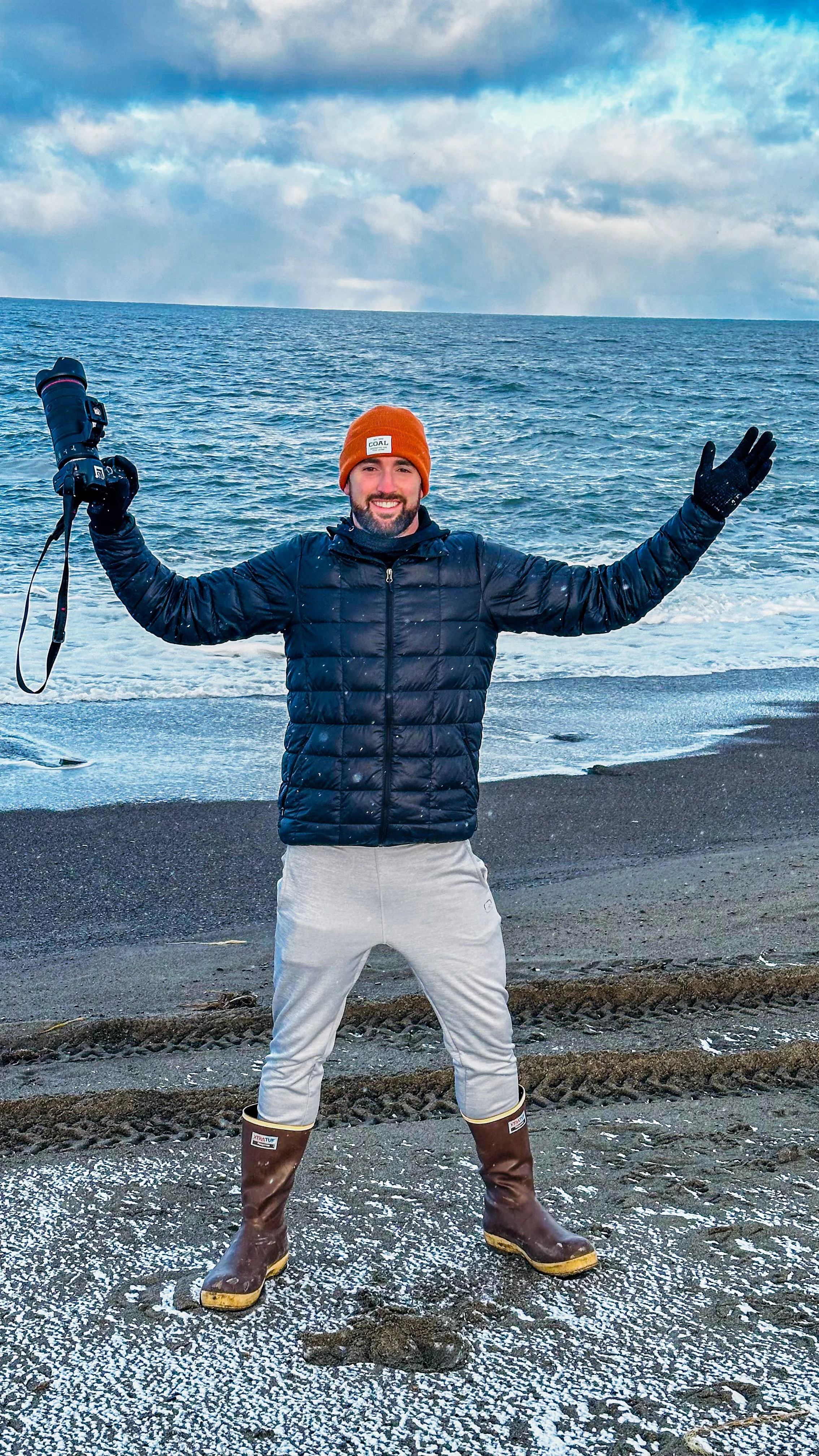 A smiling man in a black jacket, beige pants, and brown boots holding a camera, standing on a beach with ocean in the background and arms outstretched.