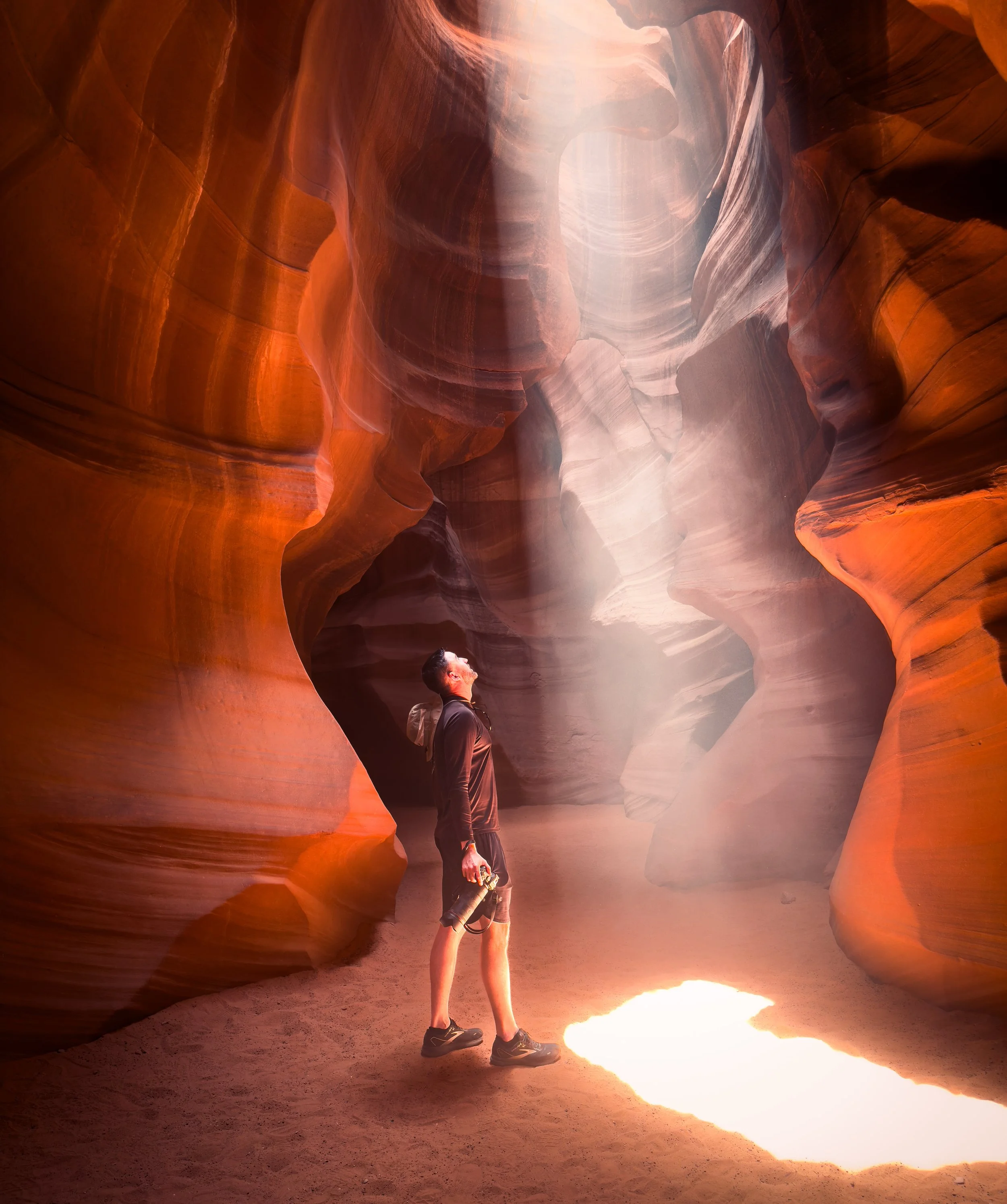 A man standing inside a slot canyon with smooth, wavy sandstone walls, looking up at a beam of sunlight coming through a narrow opening in the canyon ceiling.