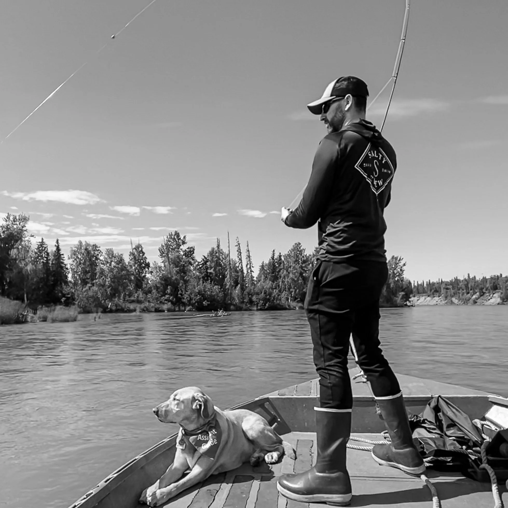 A man and a guide dog standing on a boat, fishing on a river with trees in the background, in black and white.