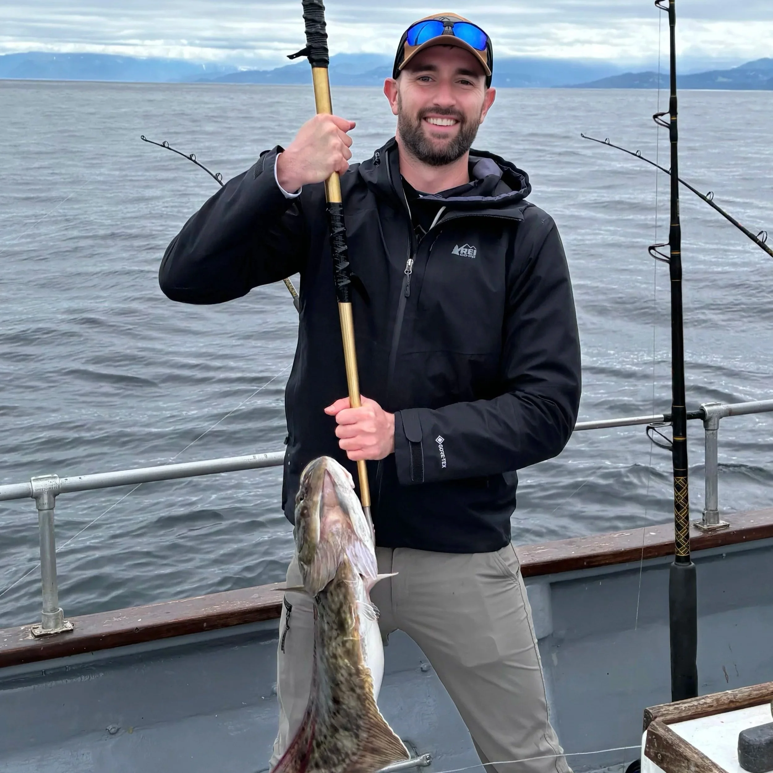 A man on a boat holding a large fish with a fishing pole, smiling at the camera, in a body of water with distant mountains in the background.
