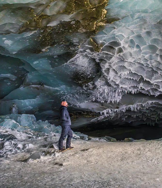 Person standing inside a frozen ice cave with blue and white ice formations covering the ceiling and walls.