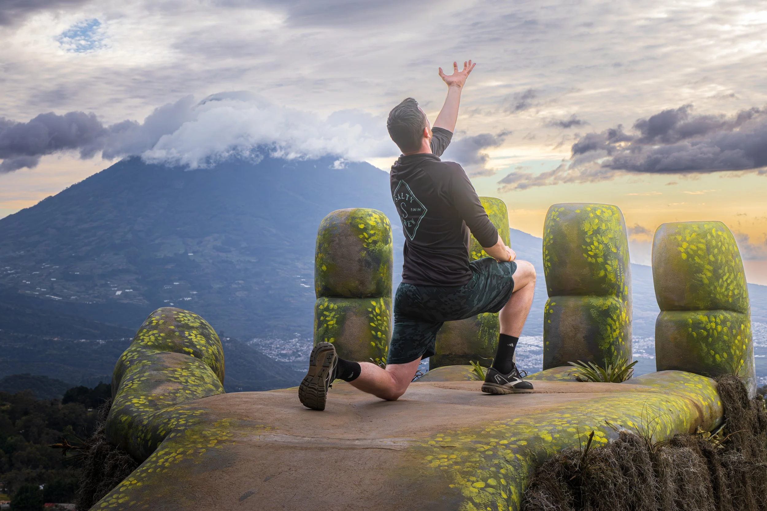 A man kneeling with one knee on the ground and one arm raised on a moss-covered stone platform with carved stone chairs, overlooking a mountainous landscape at sunset.