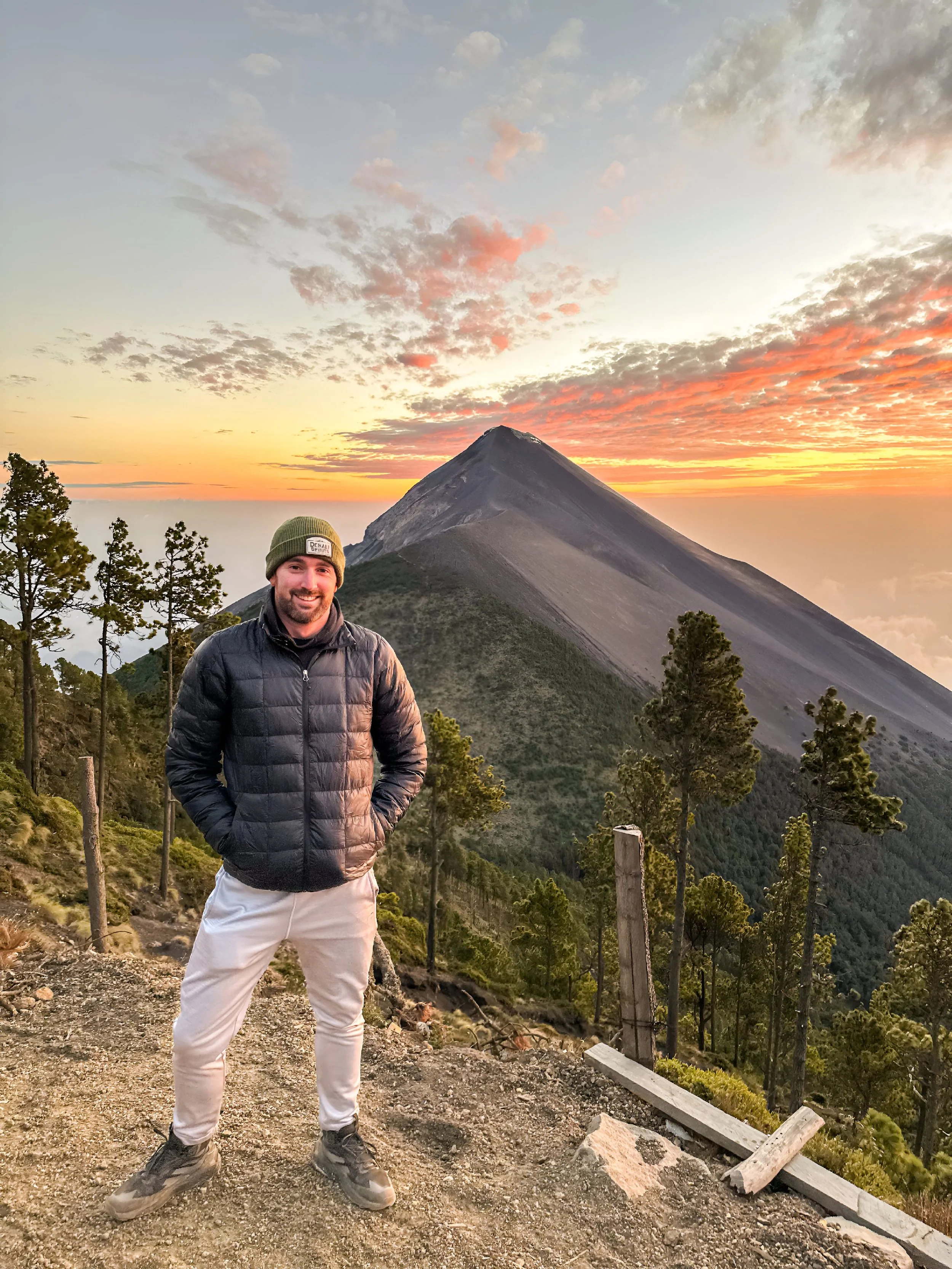 A man standing outdoors on a trail with a volcano and a colorful sunset in the background. He is smiling, wearing a black jacket, light pants, and a green beanie.