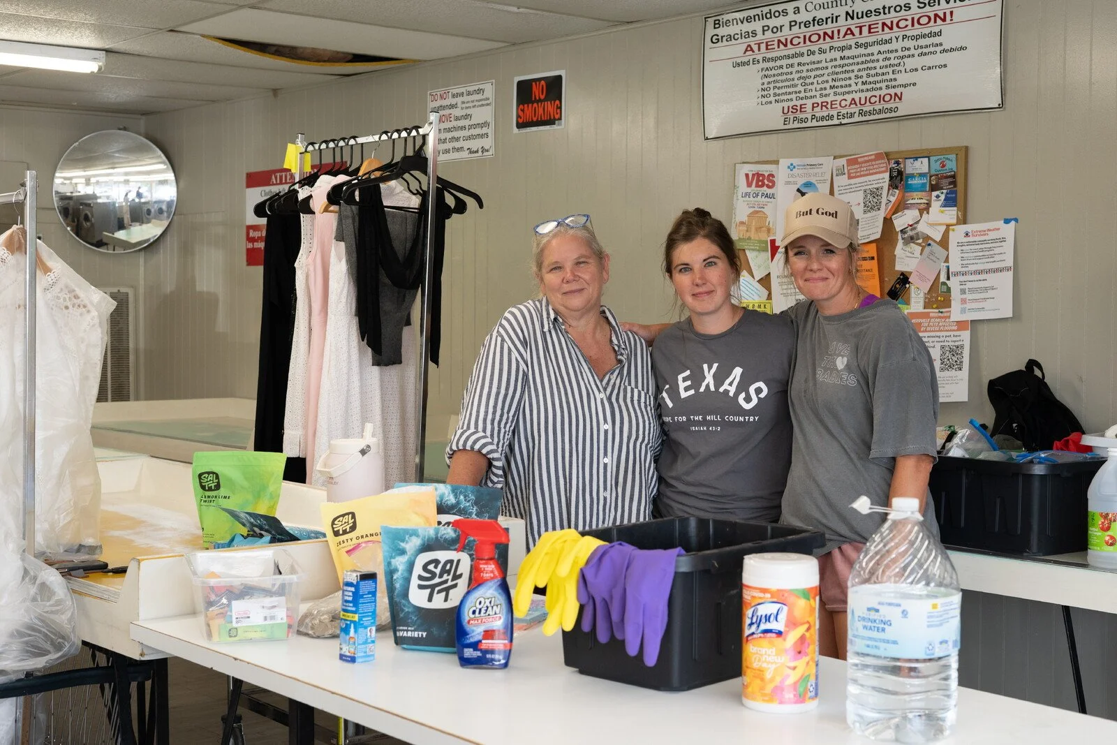 Three women standing together behind a table with cleaning supplies, wearing casual clothes, in a room with various notices and clothing items displayed.