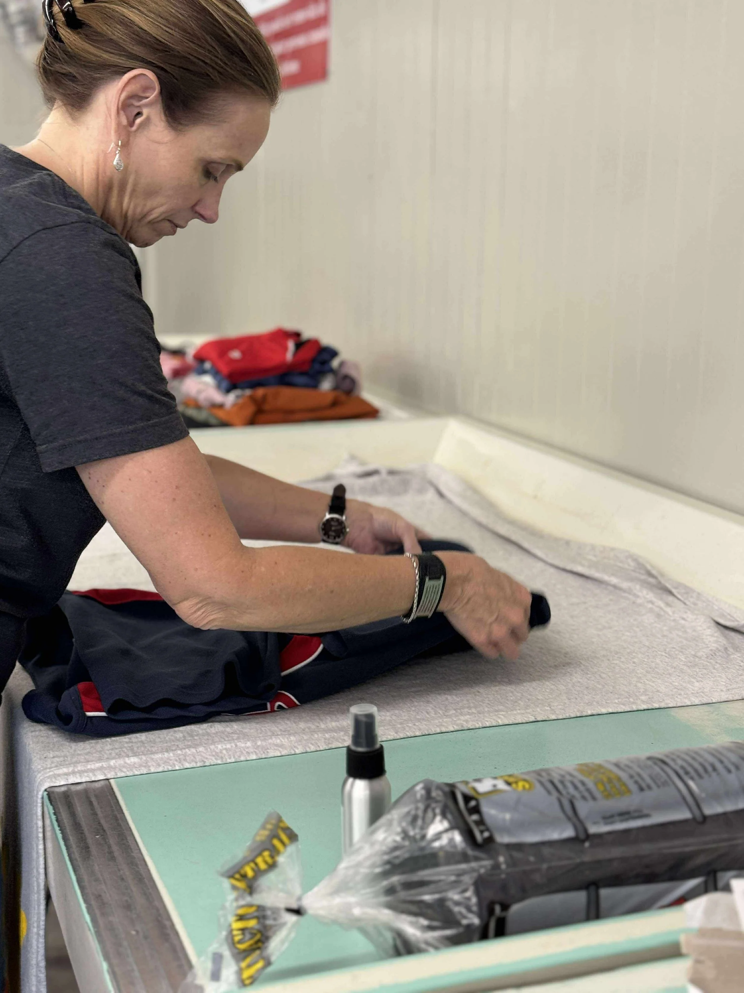 A woman folding clothes on a table in a laundry room, with laundry supplies and piles of folded clothes in the background.