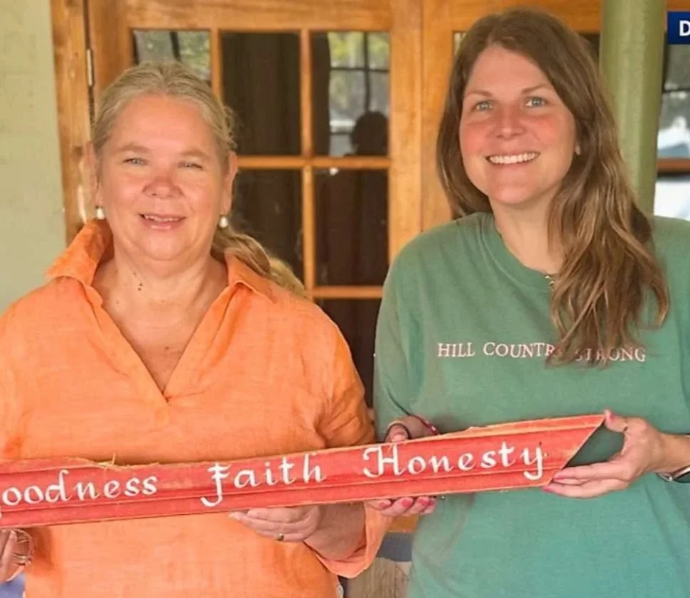 Two women indoors holding a red sign that reads 'Goodness faith Honesty'.