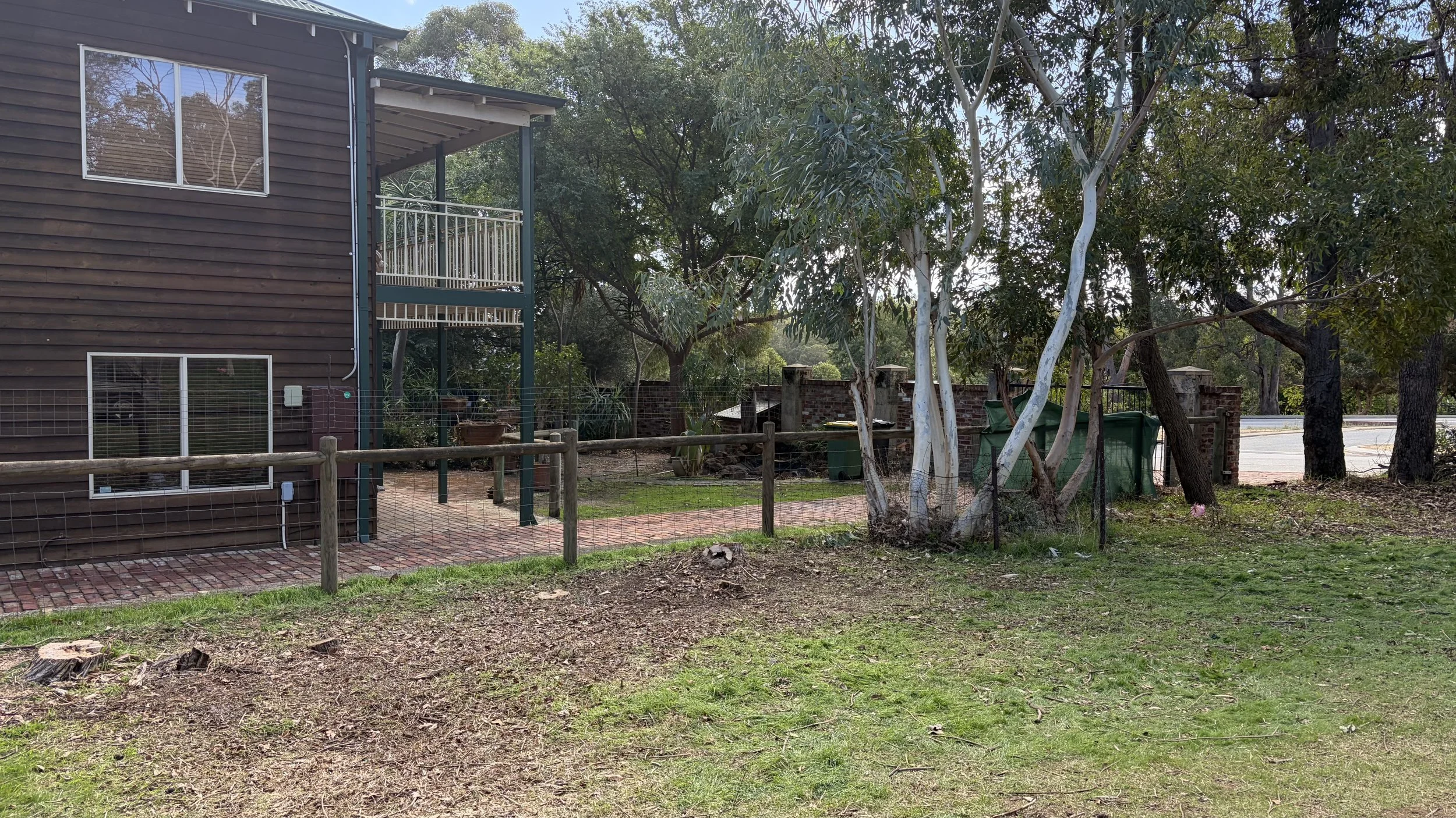 A residential backyard with a two-story house, a wraparound balcony, and trees surrounding a small fenced yard with a brick pathway and green grass.