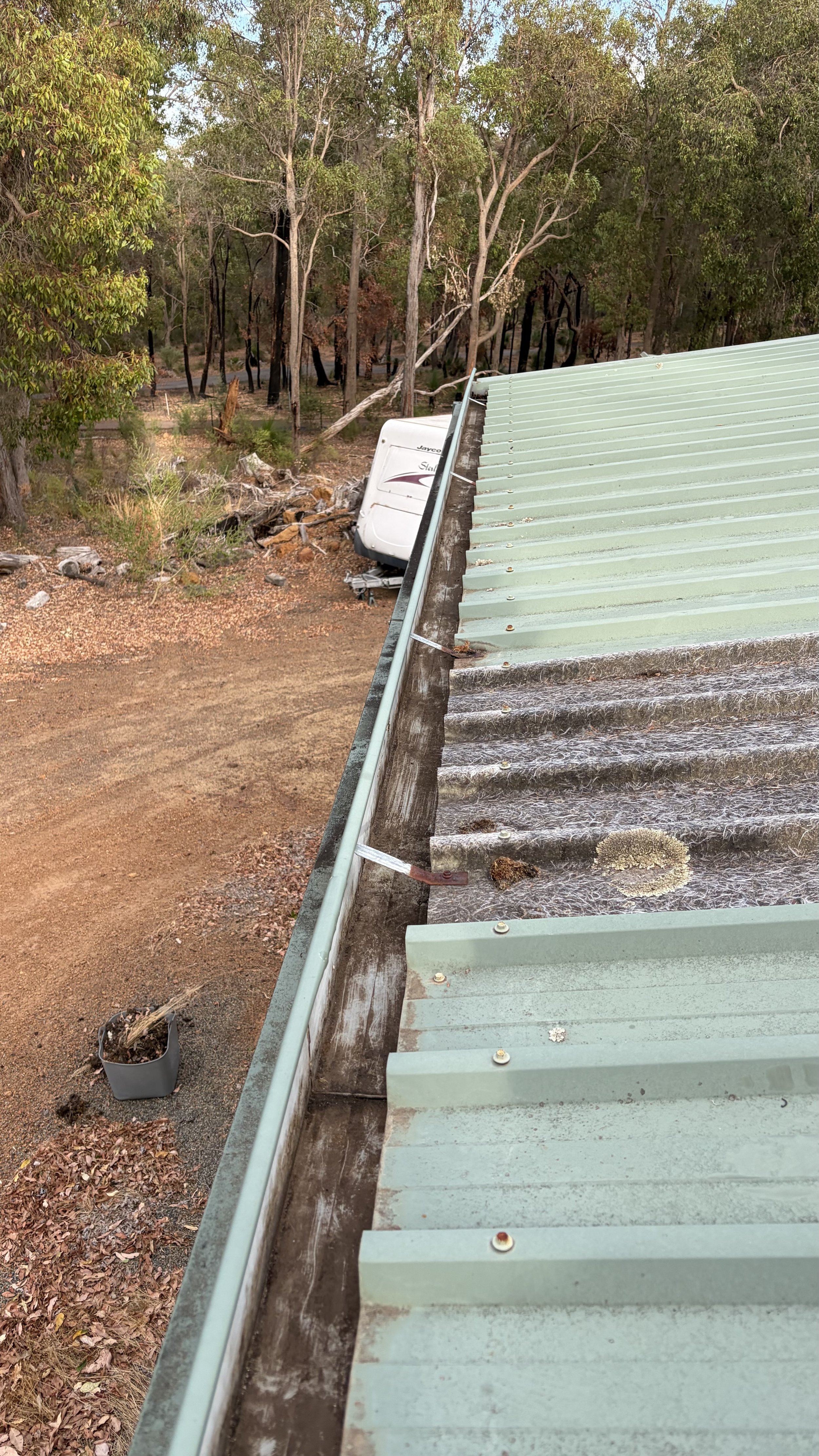 A green metal roof with some rust and moss, viewed from the edge, with a truck and wooded background.