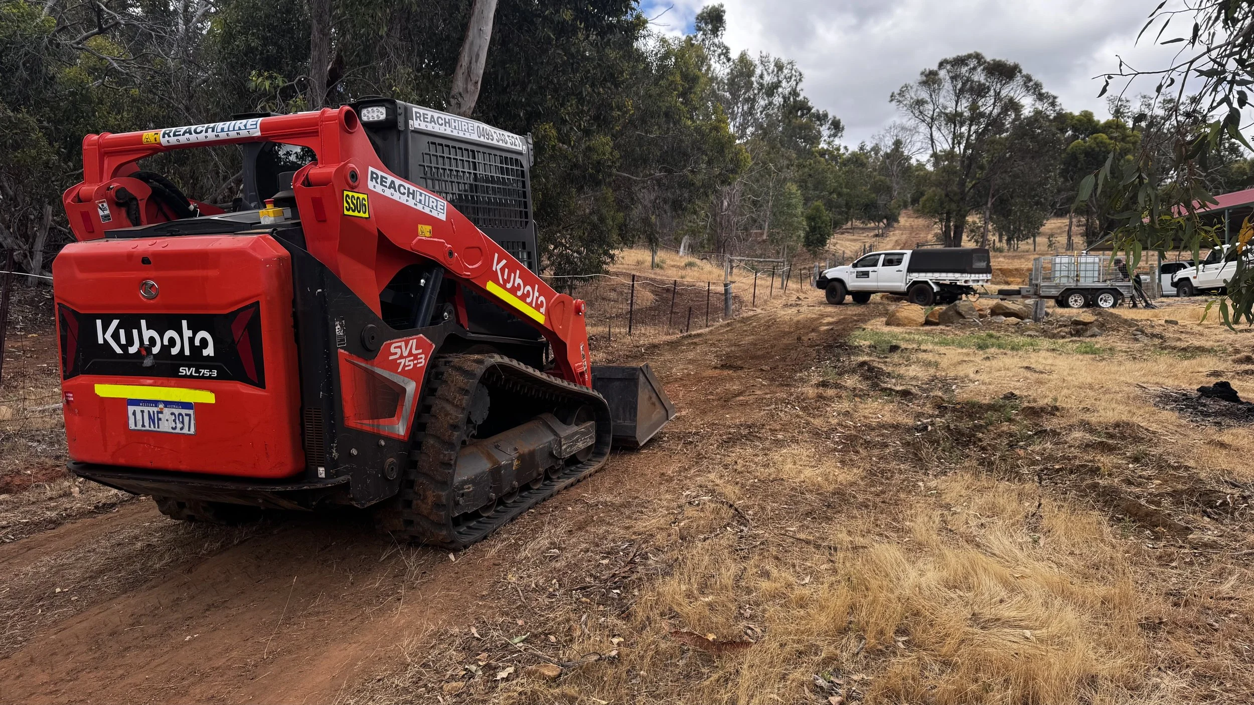 A red Kubota VL75-3 compact track loader parked on a dirt trail in a rural area with trees and dry grass, with white pickup trucks and construction equipment visible in the background.