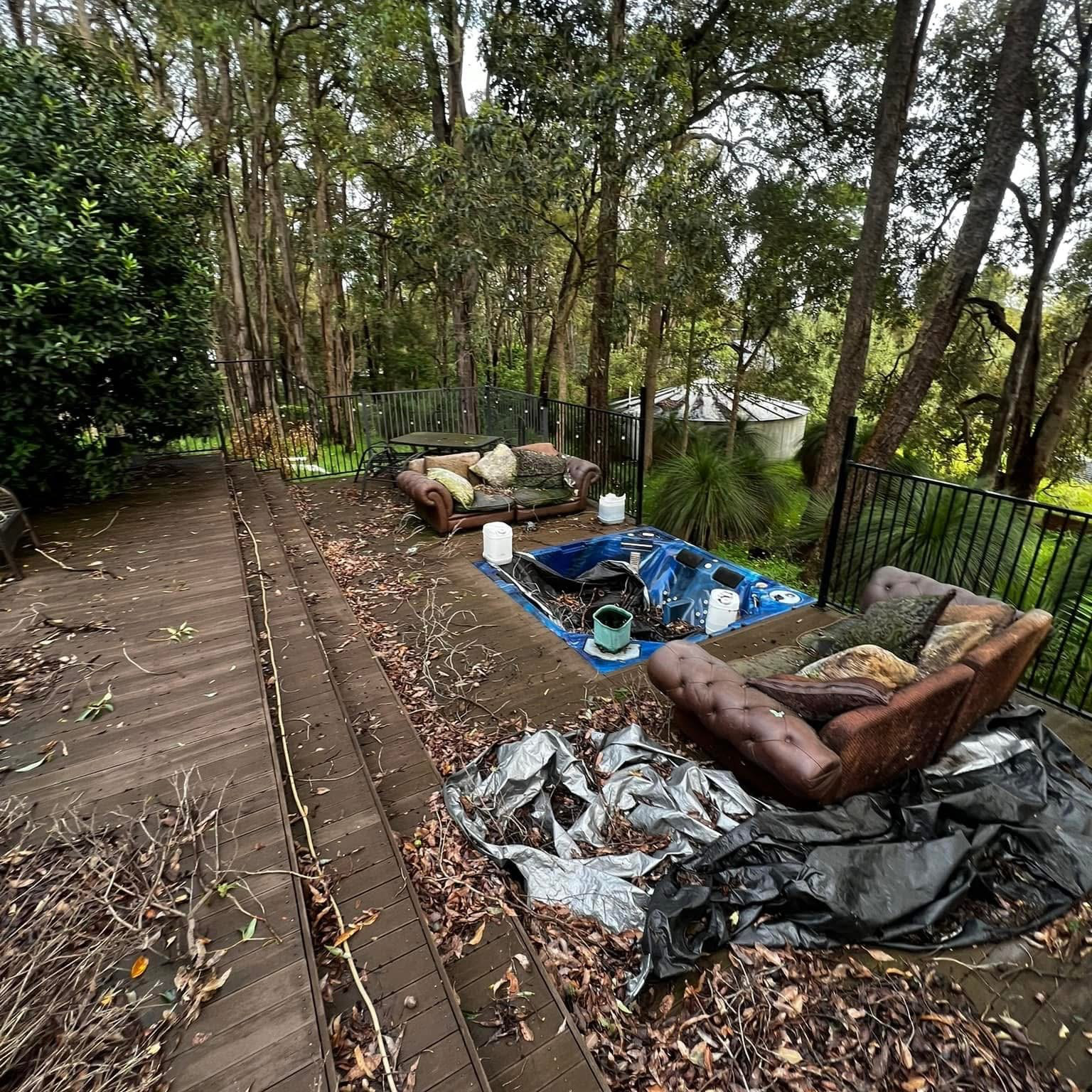 An outdoor wooden deck surrounded by trees and foliage with two sofas, a hot tub, and cleaning supplies.