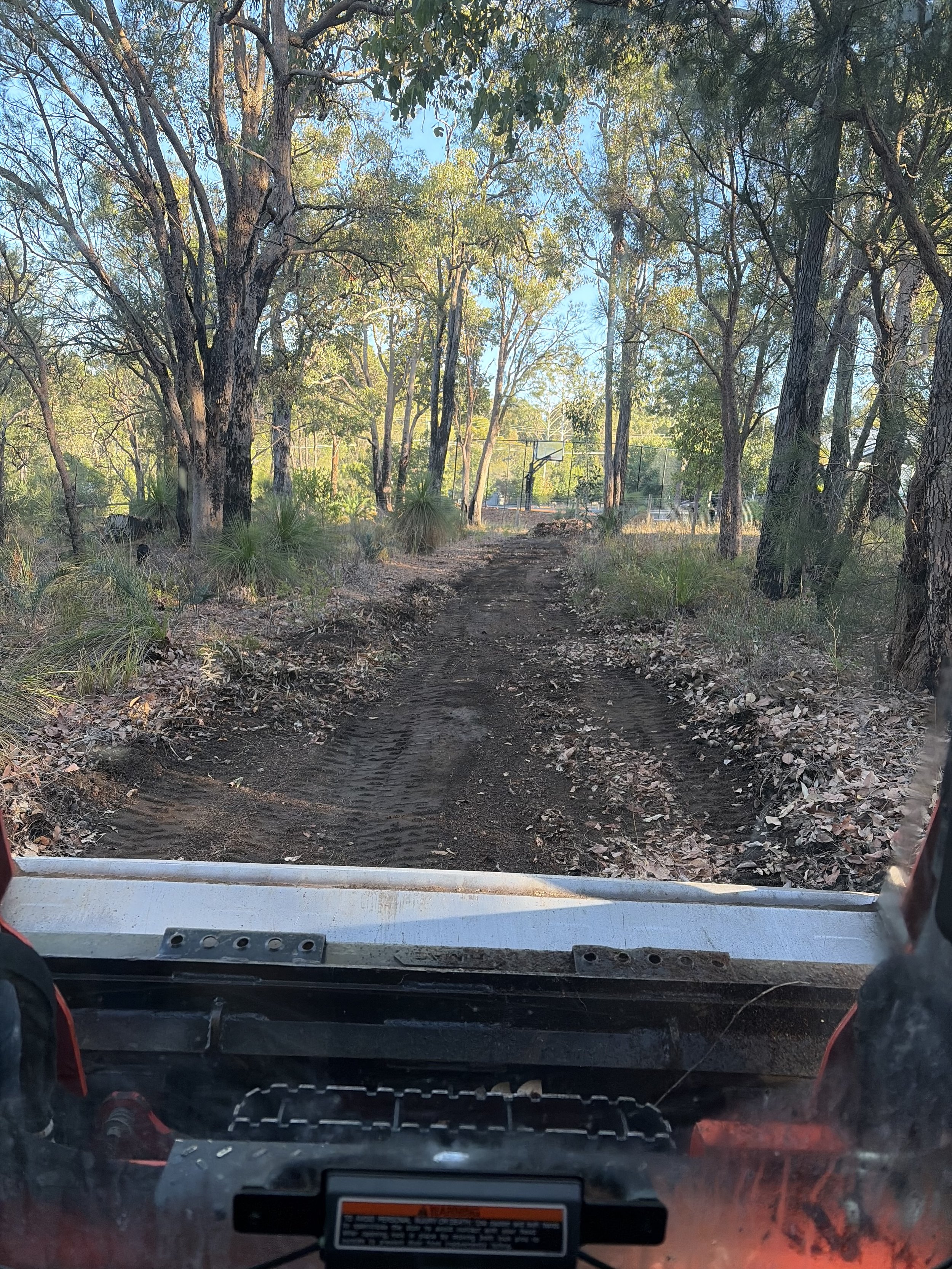 View from inside an off-road vehicle looking at a dirt trail through a wooded area with trees and a basketball court in the background.