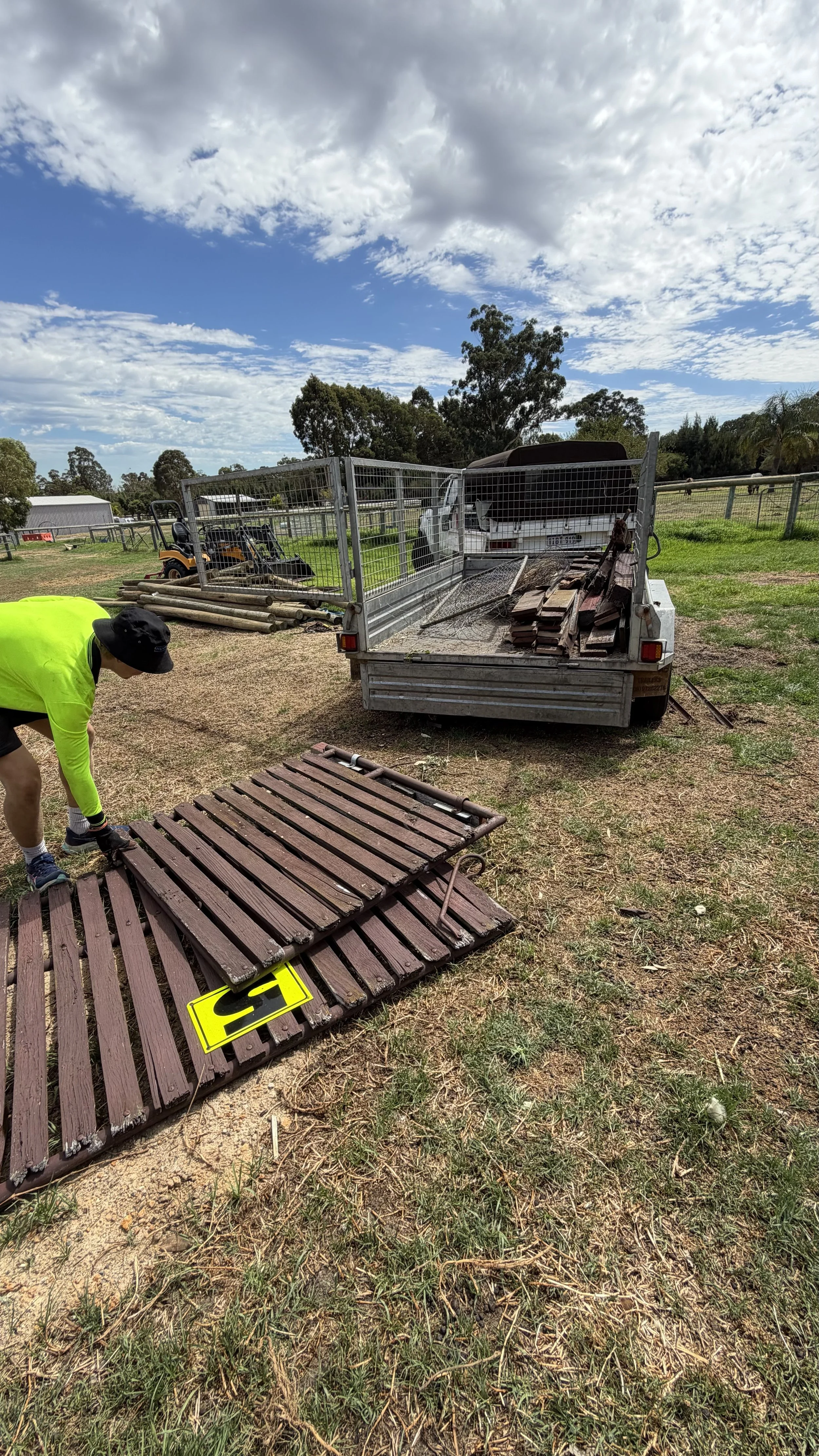 A worker in a neon yellow shirt and black hat is lifting a brown wooden bench panel onto the ground in a rural area with trees and a partly cloudy sky. There is a trailer with more wooden panels and a white vehicle in the background.
