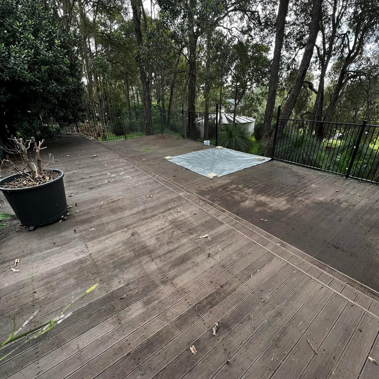 Empty wooden deck with a black metal fence, potted plant, and a plastic sheet on the ground, surrounded by trees and greenery.