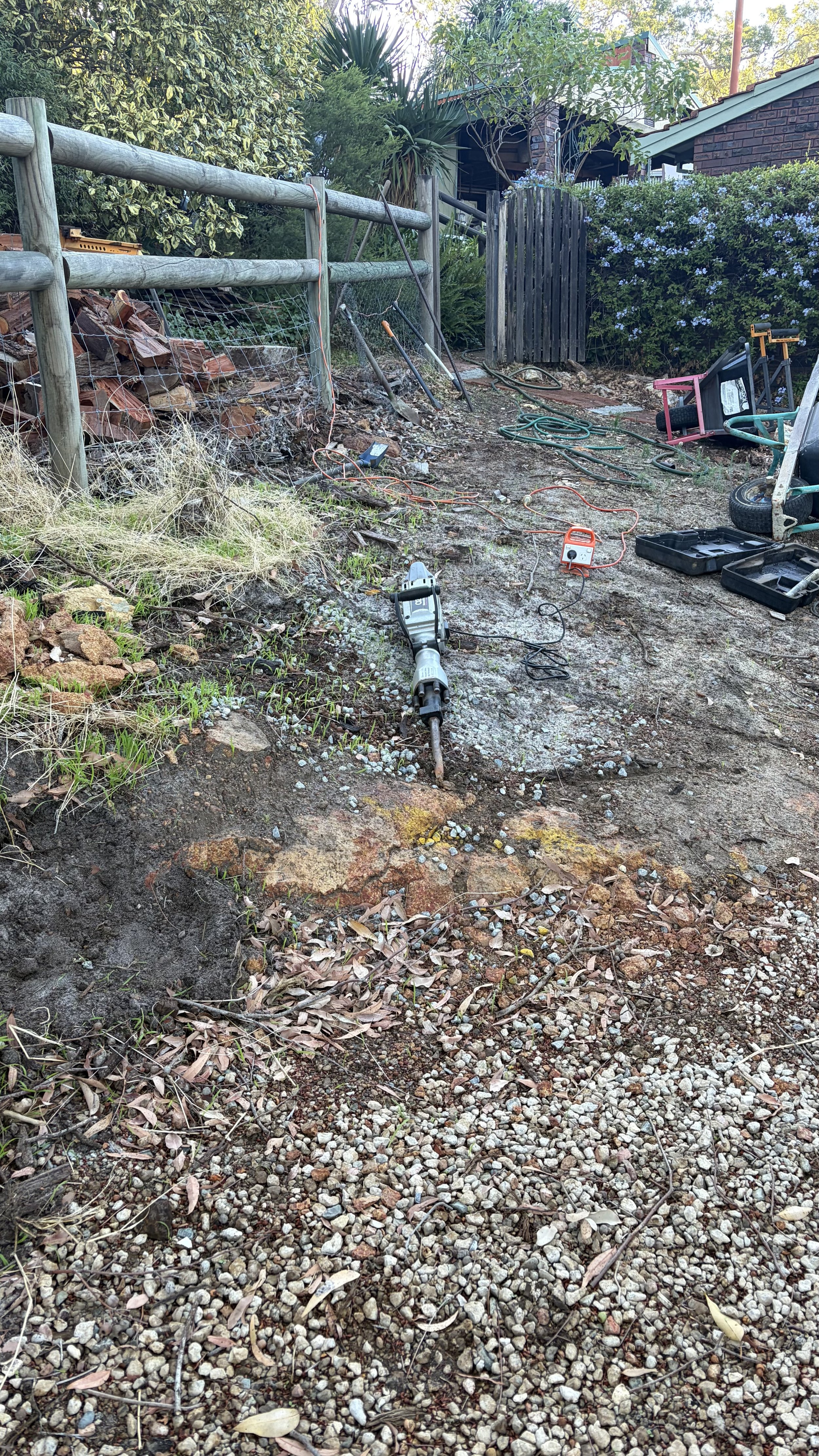 A backyard garden area under construction with power tools, extension cords, and construction materials scattered on the ground. There is a grassy and gravel surface, a wooden fence, and an open door leading to a house in the background.