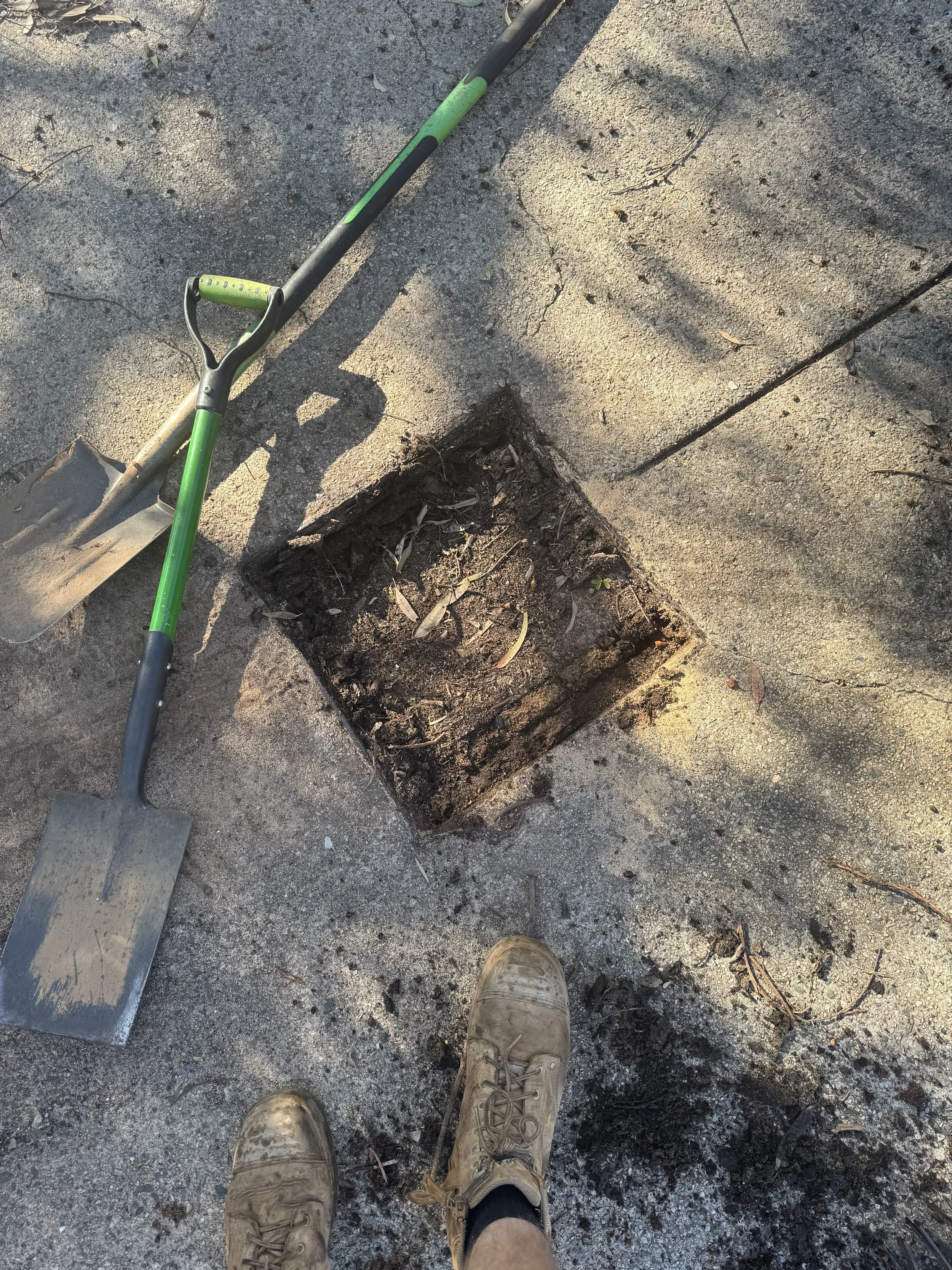 Worker's muddy boots stand on a cracked sidewalk next to a square hole in the ground, with a shovel resting nearby.