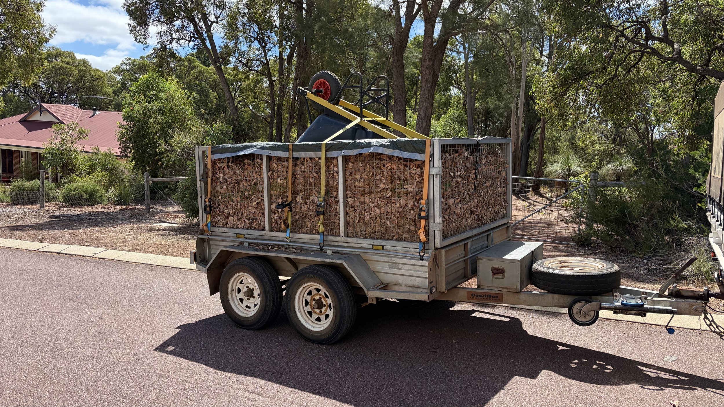 A small utility trailer loaded with chopped firewood and secured with straps, attached to a vehicle, parked on a paved street with trees and a house with a red roof in the background.