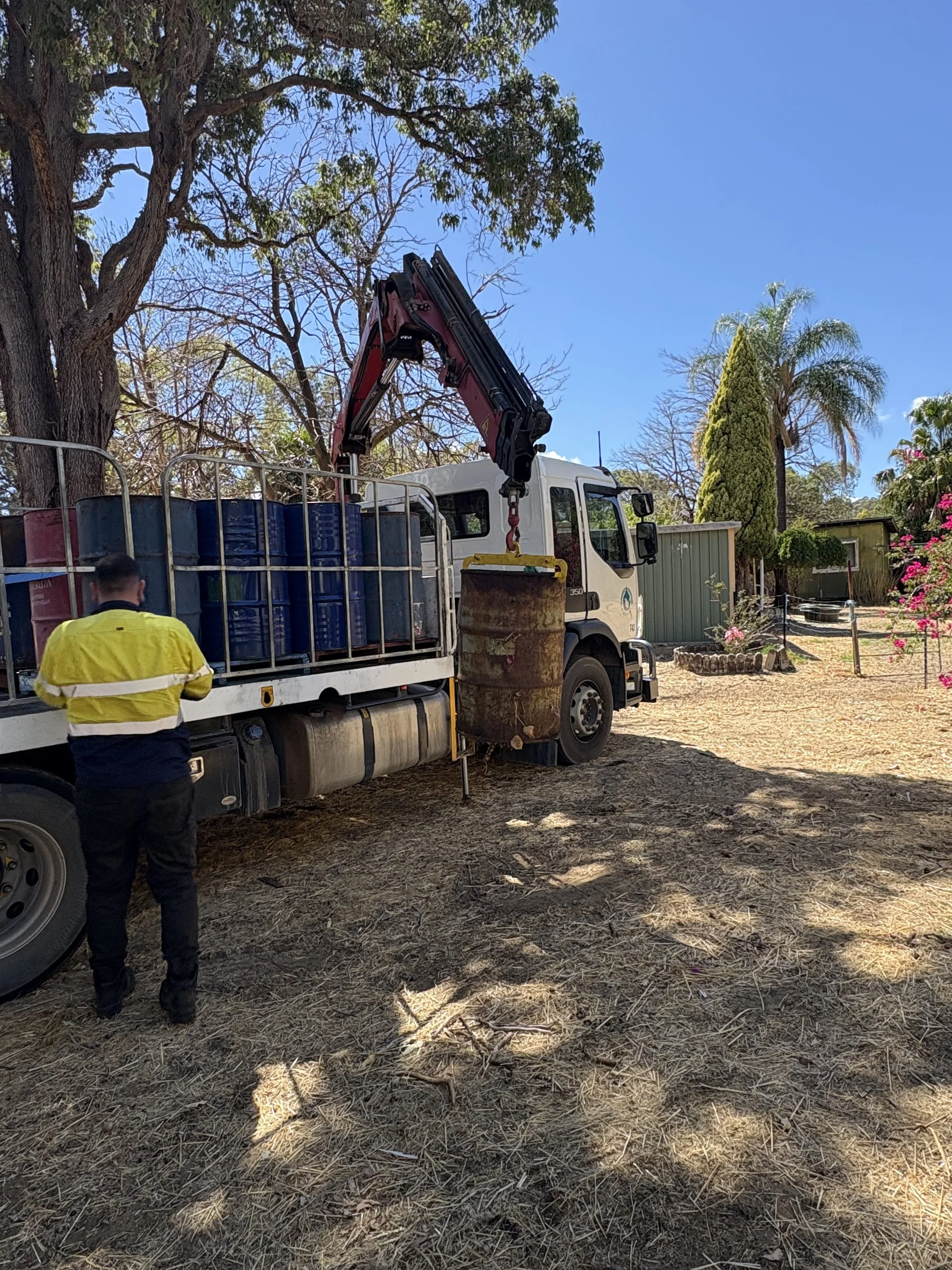 A worker standing next to a truck with a crane lifting a rusted metal barrel in a backyard with trees and flowers.