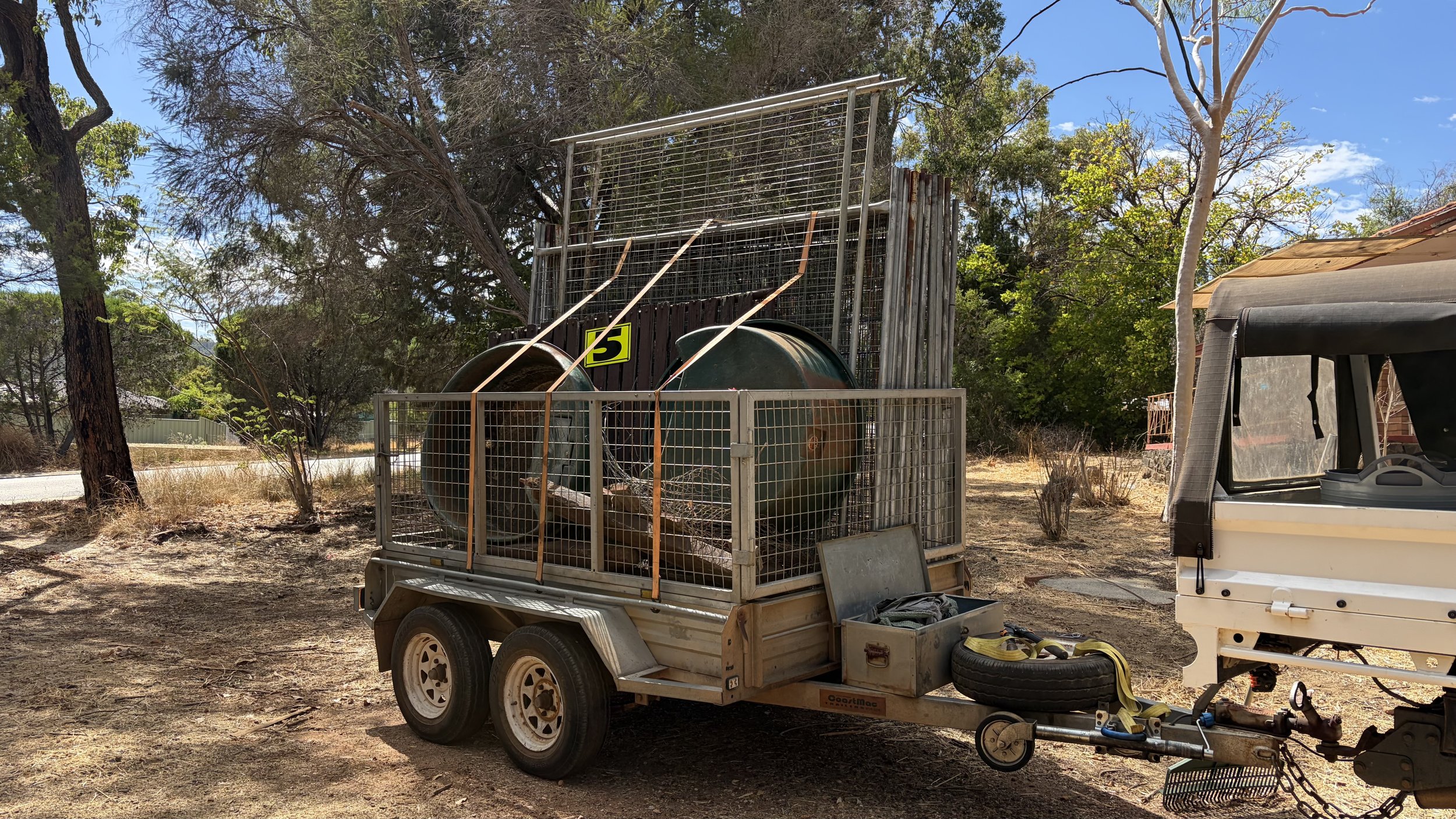 A small trailer with two large metal tanks, a tool box, and a spare tire, parked outdoors with trees and clear blue sky in the background.